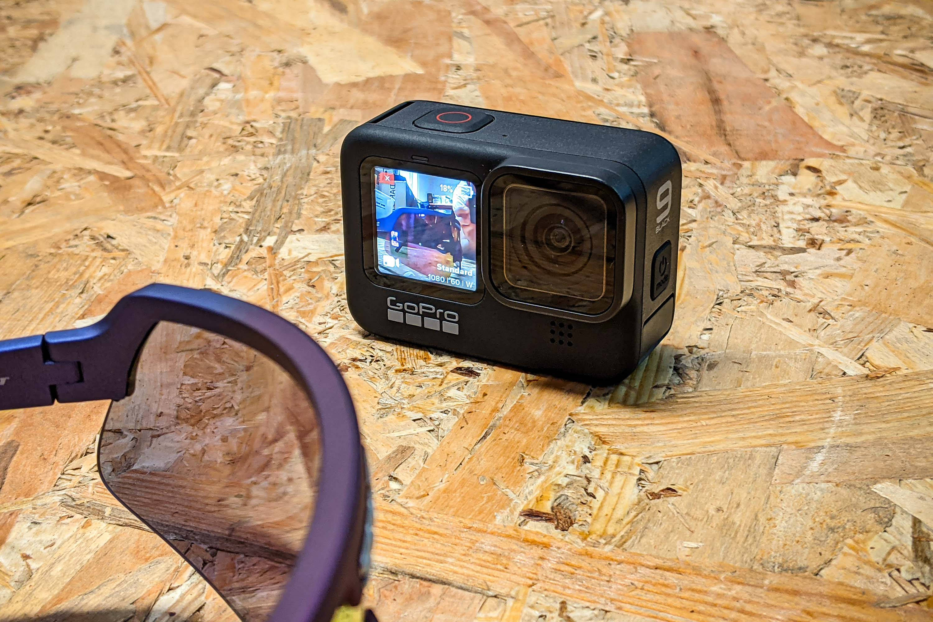 攝影 桌布 壁紙 — 一台黑色GoPro 9相機放在有紋理的OSB木板上，旁邊是紫色運動眼鏡，在明亮的日光下。