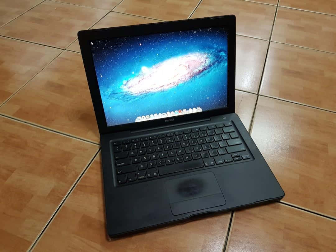 Black Macbook 桌布 壁紙 — 一台黑色MacBook筆記型電腦打開放在木紋磚地上，顯示出充滿活力的螺旋星系桌布。