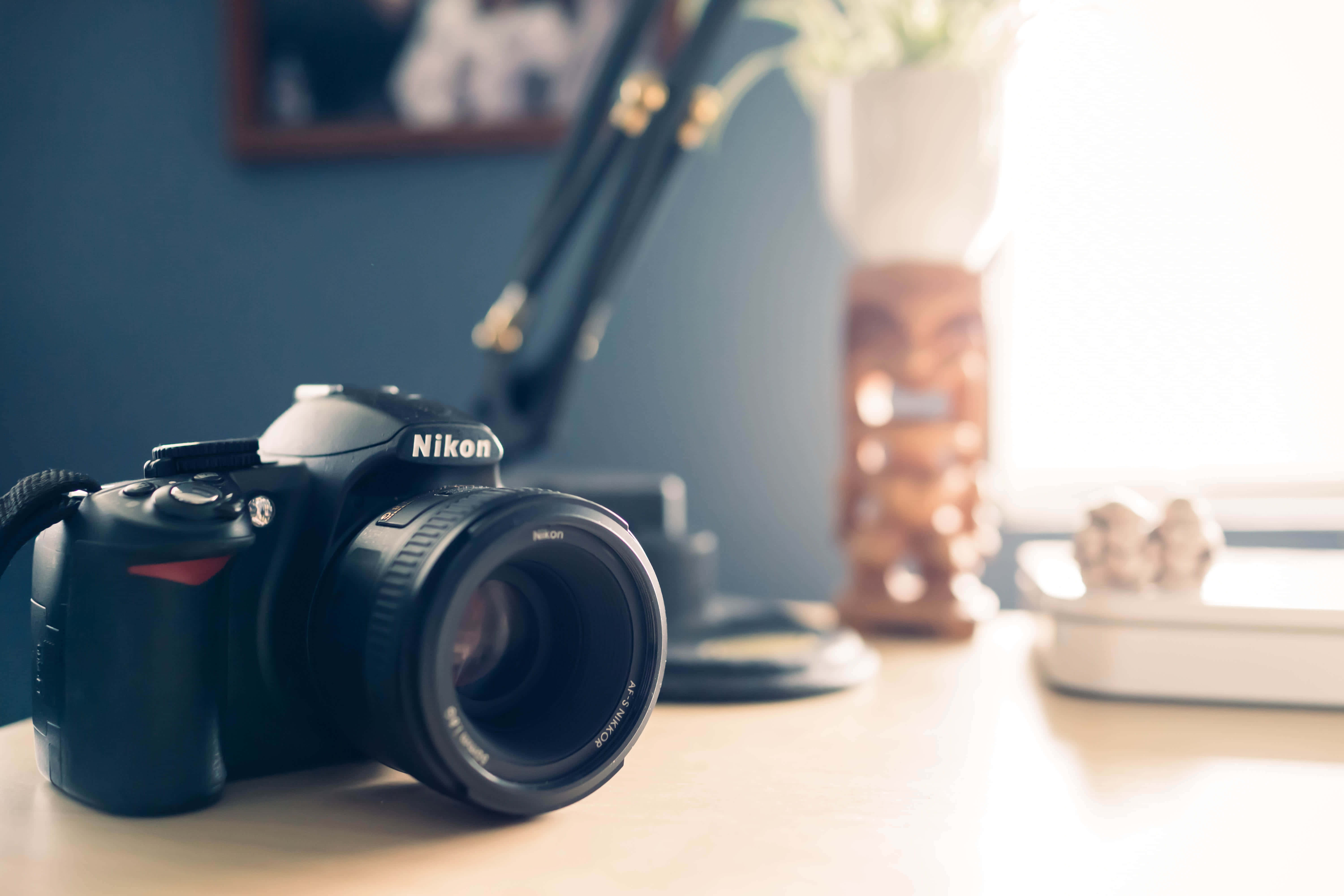 Dslr 桌布 壁紙 — 一台黑色Nikon DSLR相機放在木桌上，柔和自然光從裝飾花瓶後的窗戶射入。