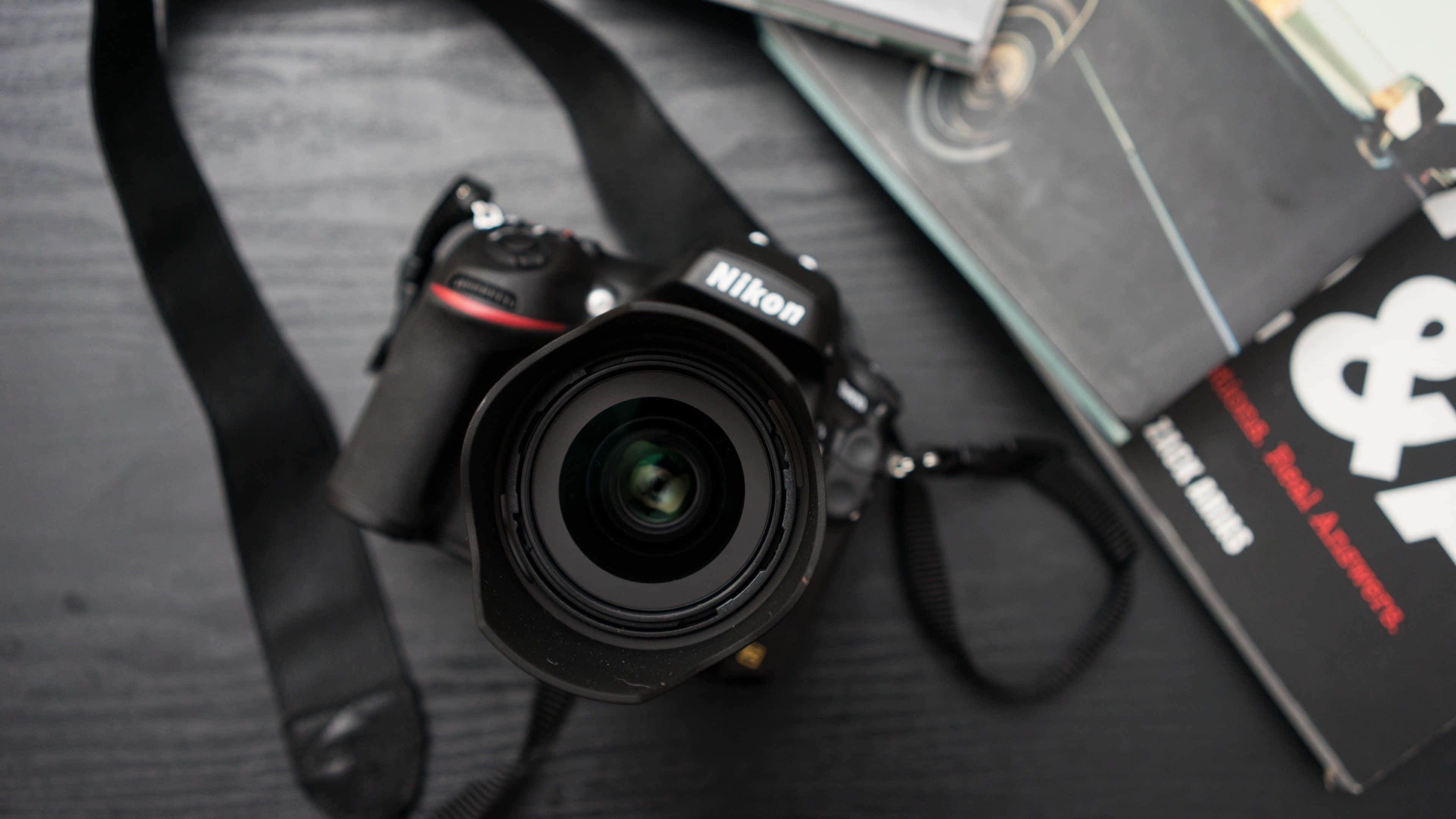 攝影 桌布 壁紙 — 一台黑色Nikon DSLR相機連鏡頭放置在深色木質表面上，旁邊有攝影書籍。