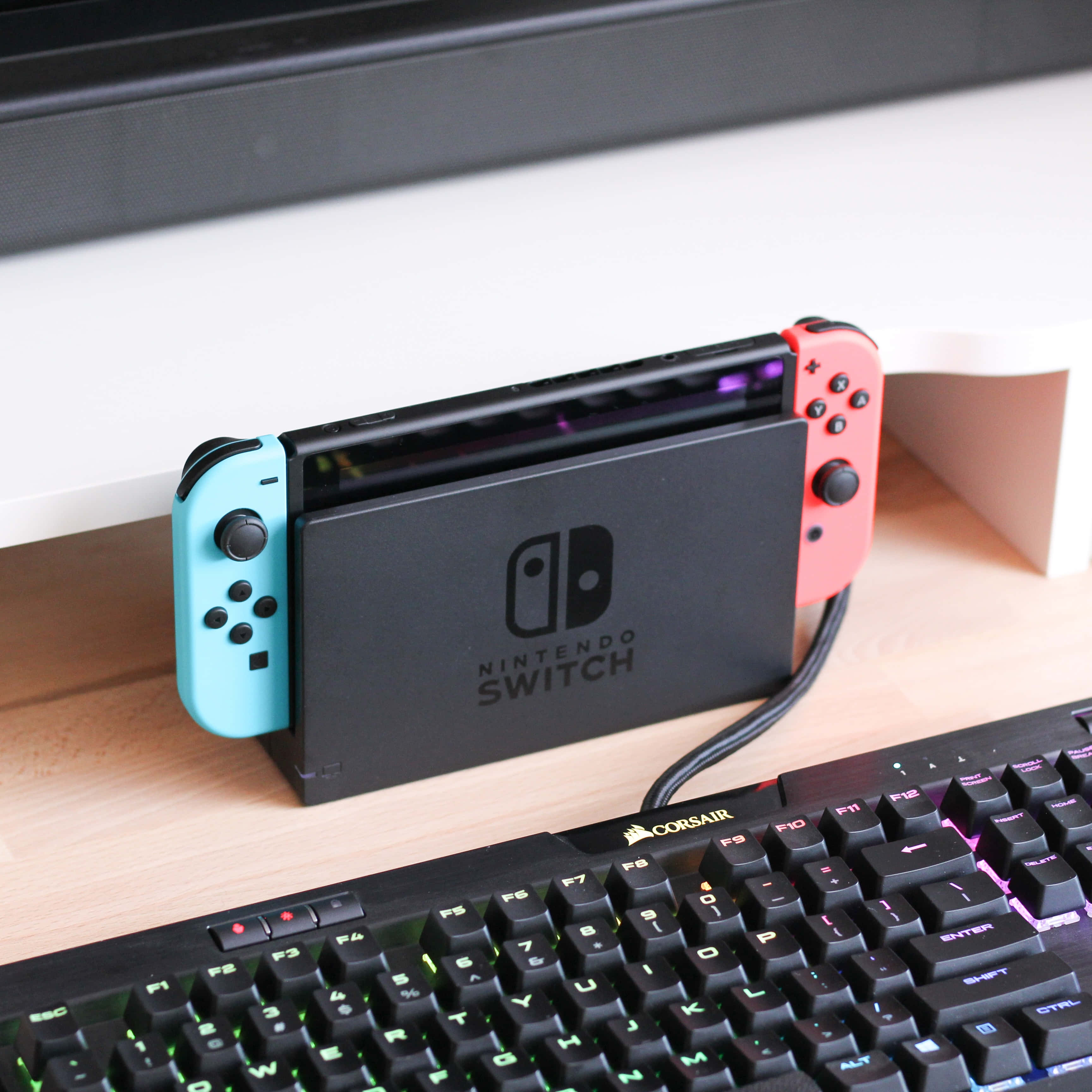 Gaming Consoles 桌布 壁紙 — 一台黑色Nintendo Switch配備綠松色和珊瑚色Joy-Con，直立放在木桌上，旁邊有一個發光的Corsair鍵盤。