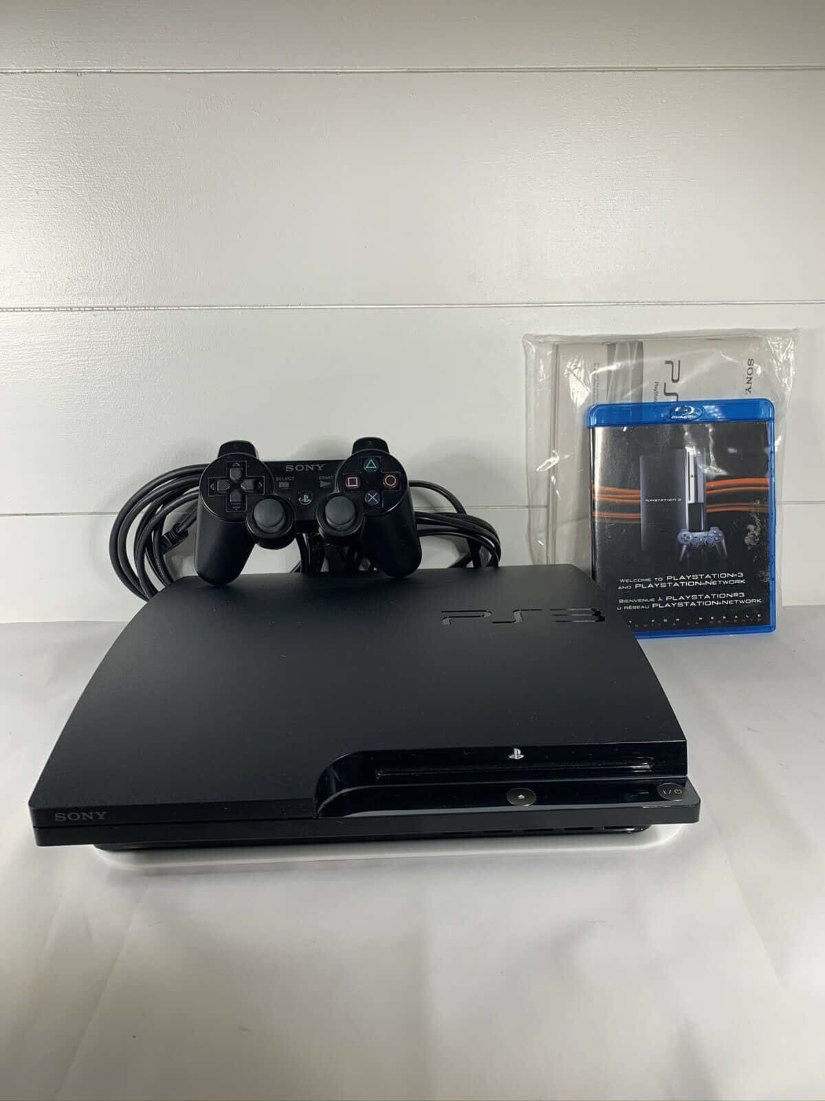 PlayStation 桌布 壁紙 — 一台黑色PlayStation 3主機、手把及遊戲盒放在白色表面上，背景是白色板牆。