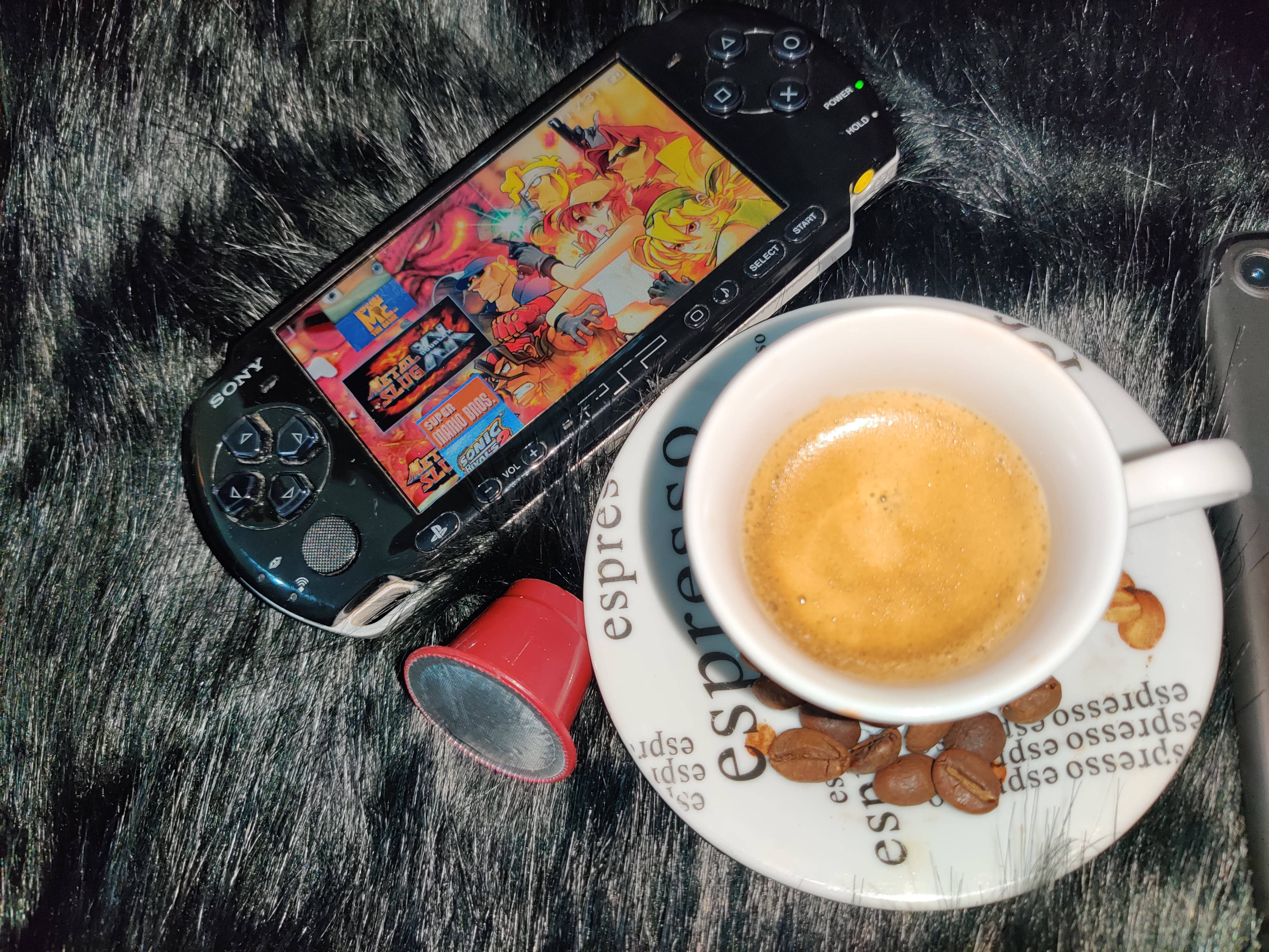 PlayStation 桌布 壁紙 — 一台黑色PSP正在玩色彩繽紛的動畫遊戲，旁邊放著一杯冒煙的濃縮咖啡，置於毛絨黑色表面上。