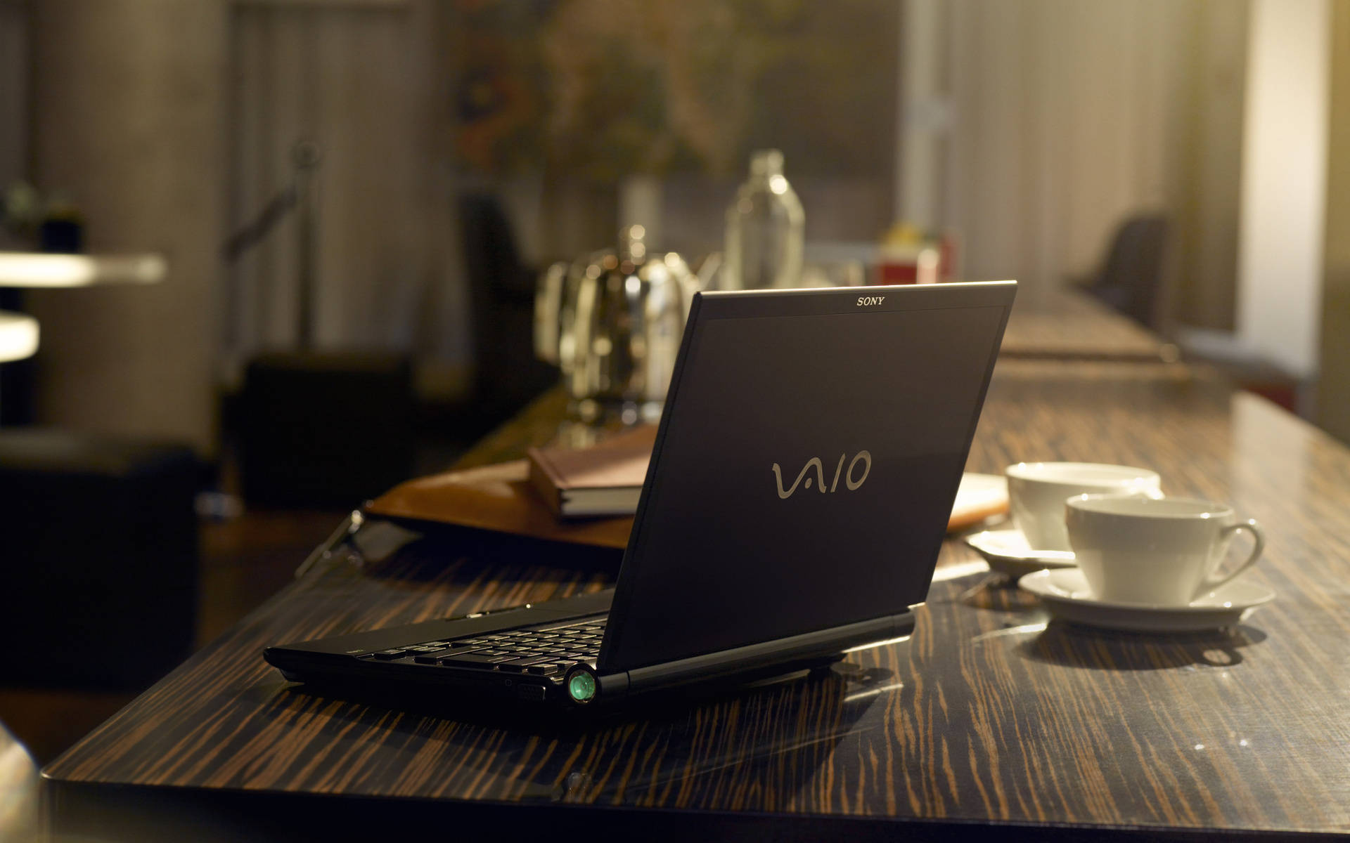 Sony 桌布 壁紙 — 一台黑色Sony VAIO筆記型電腦打開放在深色木桌上，旁邊有兩個白色咖啡杯，在一個燈光溫暖的咖啡館裡。