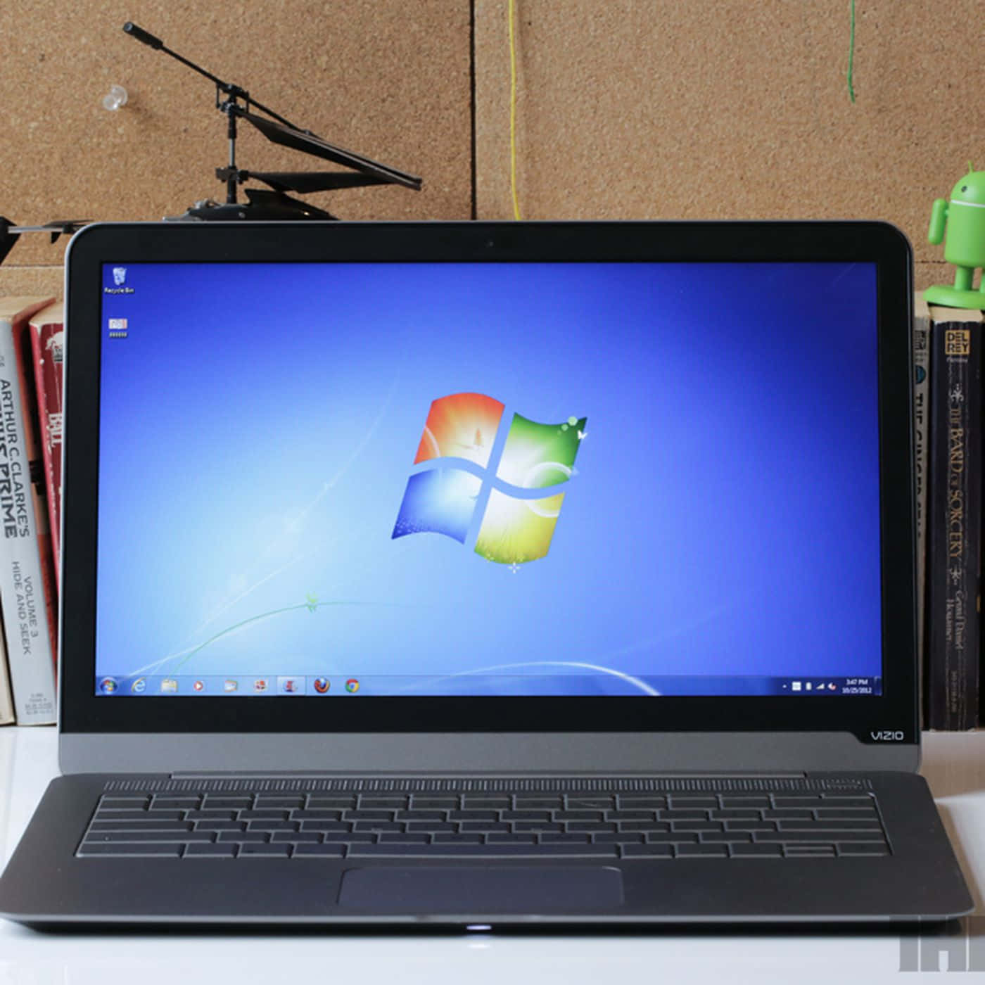 Windows 7 桌布 壁紙 — 一台黑色Vizio筆記型電腦在桌上顯示經典的Windows 7桌布，旁邊有書本和一個綠色Android公仔。