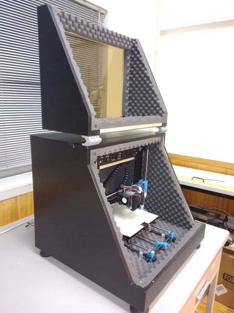 3d Printer 桌布 壁紙 — 一台黑色帶藍色組件的3D打印機，放置在白色書桌上靠近有百葉窗的窗戶，位於一個定制的泡棉內襯外殼內。
