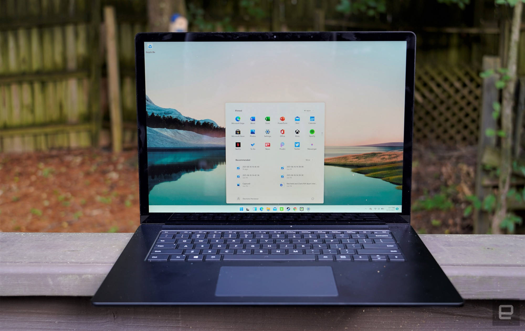 Windows 11 桌布 壁紙 — 一台黑色筆記型電腦打開放在木長凳上，顯示寧靜的山湖桌布，上面覆蓋著應用程式圖示。