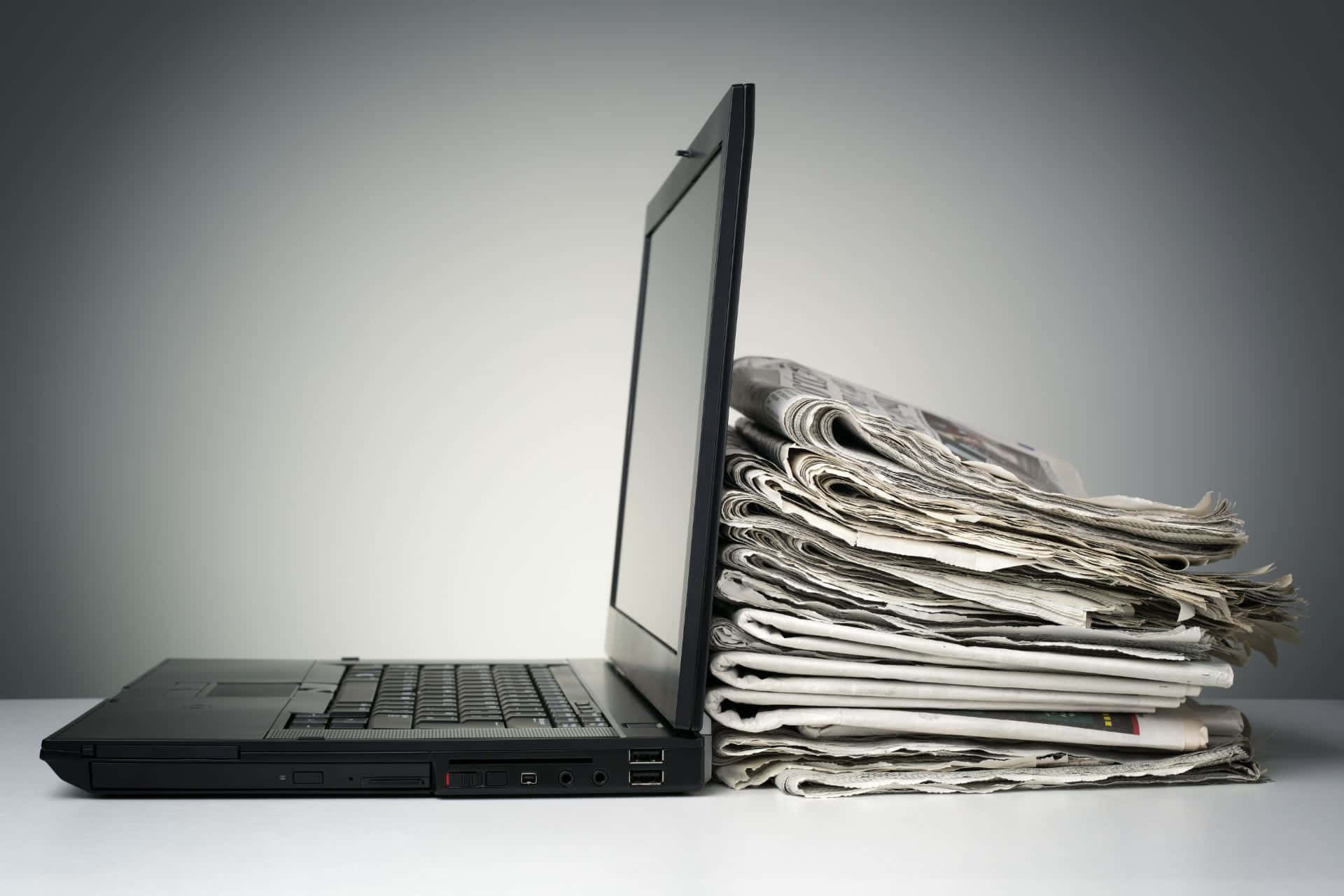 Journalism 桌布 壁紙 — 一台黑色筆記型電腦直立靠在一疊折疊報紙上，置於白色表面上。