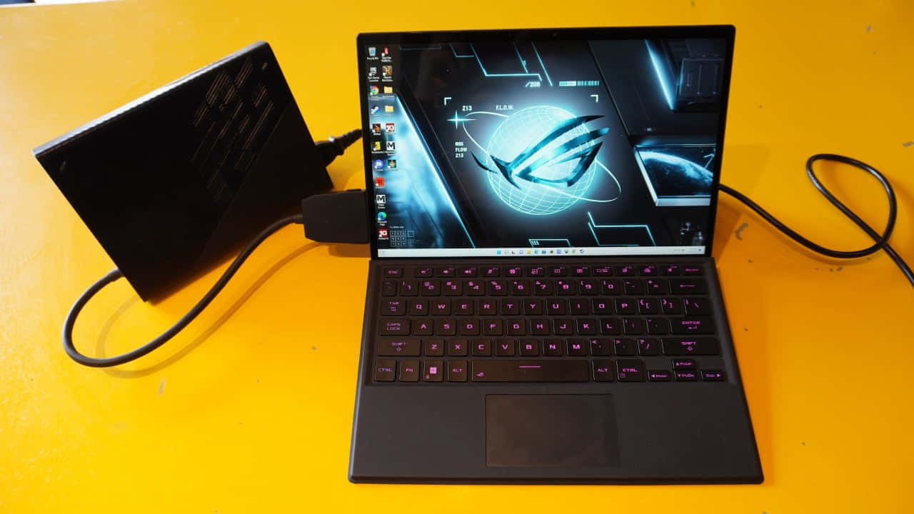 華碩 ROG 桌布 壁紙 — 一台黑色電競筆記型電腦，帶有發光紫色鍵盤，打開在亮黃色表面上，顯示未來風格的藍色和黑色桌布。