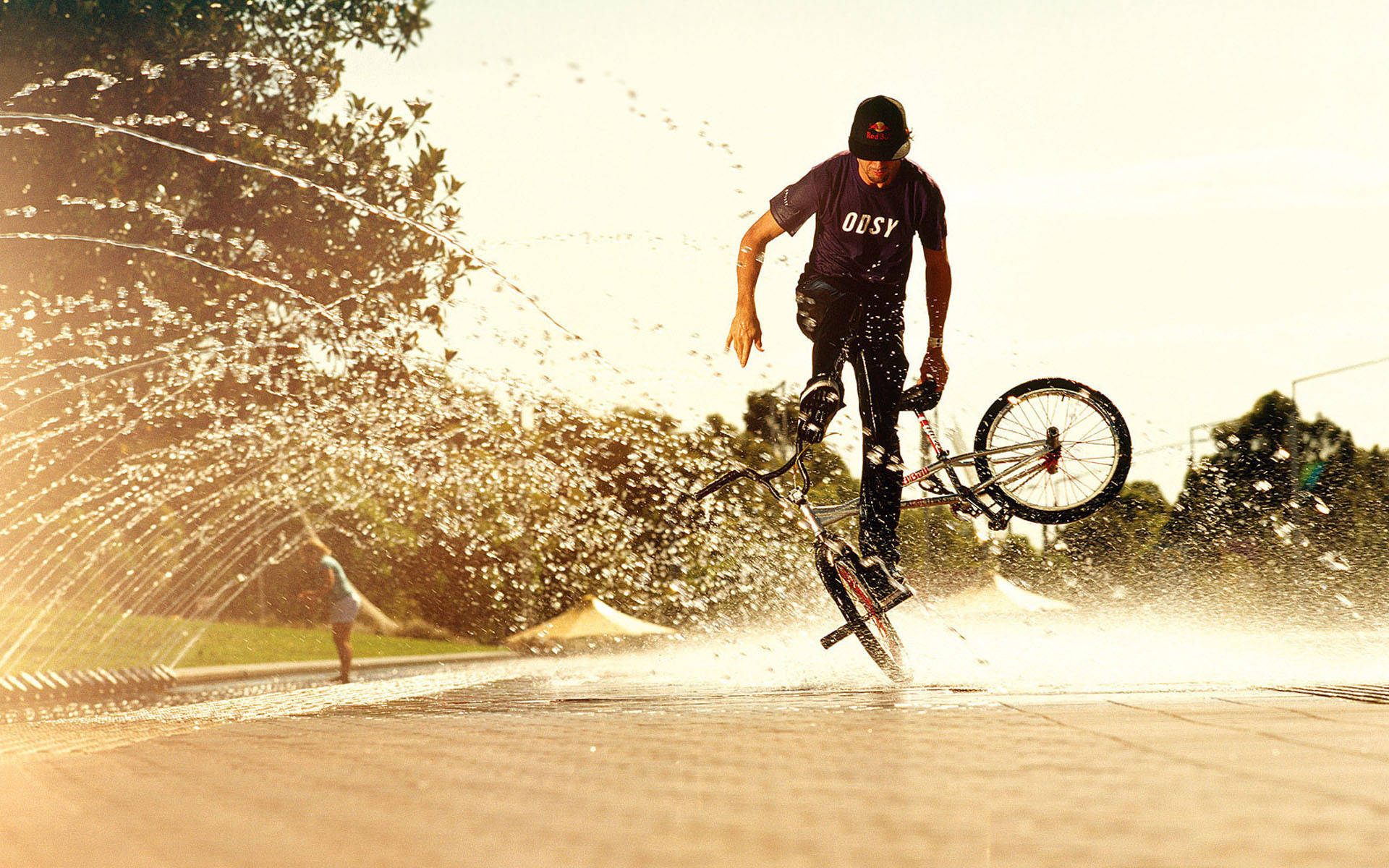 Man 桌布 壁紙 — 一名BMX車手在陽光明媚的公園中，在噴灑金色水花的噴泉上方表演空中特技。