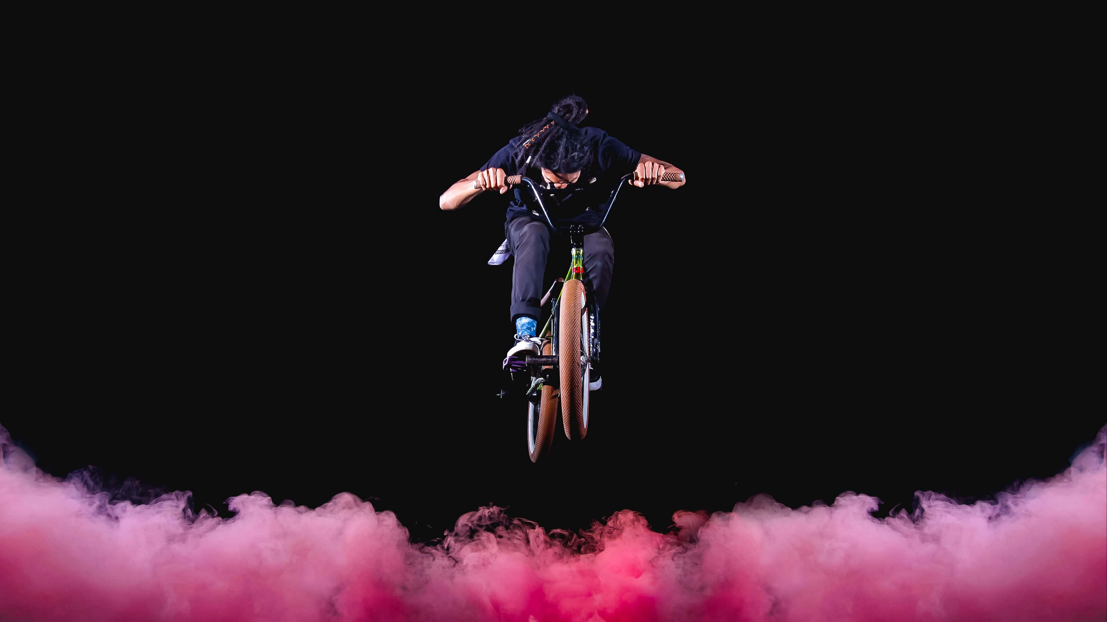 4K Bikes 桌布 壁紙 — 一名bmx車手在鮮豔的粉紅色煙霧雲上方騰空而起，背景是純黑色。