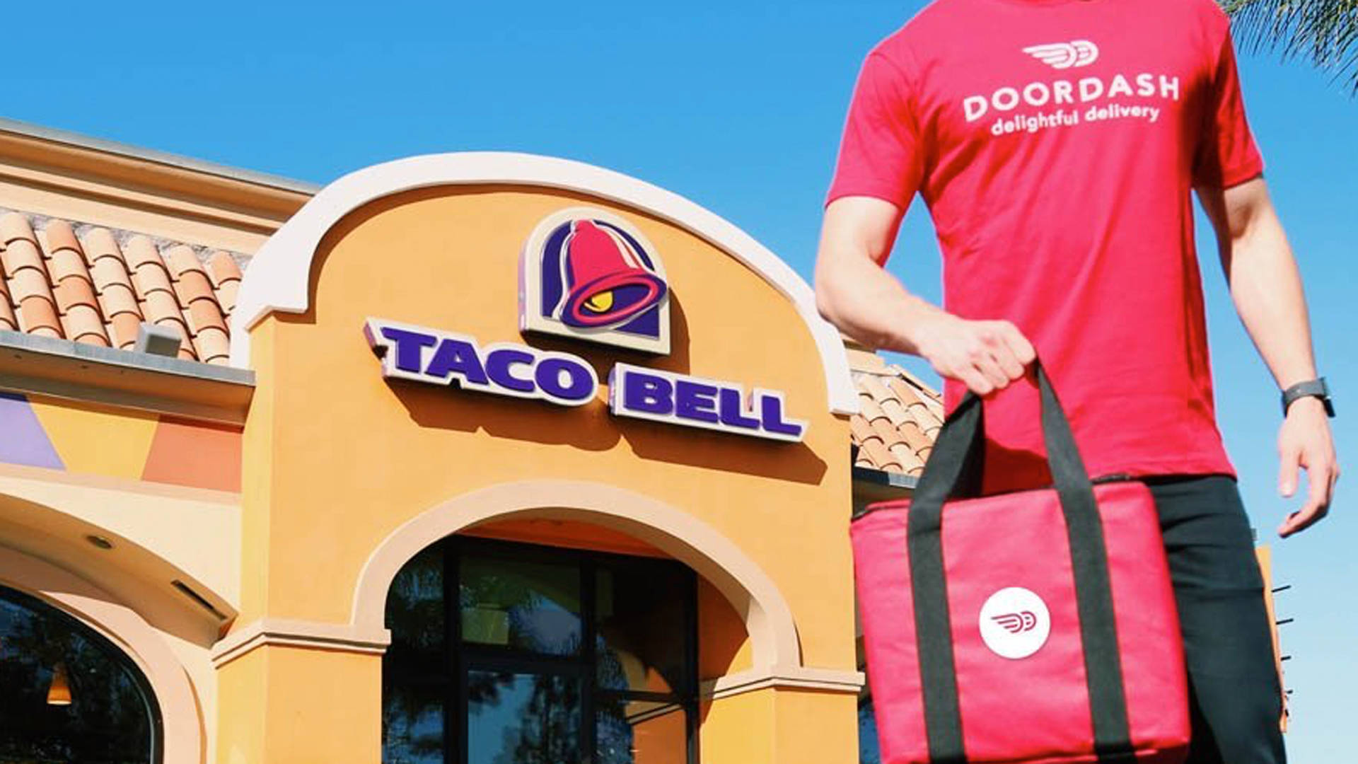 塔可鐘 桌布 壁紙 — 一名Doordash送貨員站在Taco Bell餐廳外，在晴朗的藍天下拿著一個紅色保溫袋。