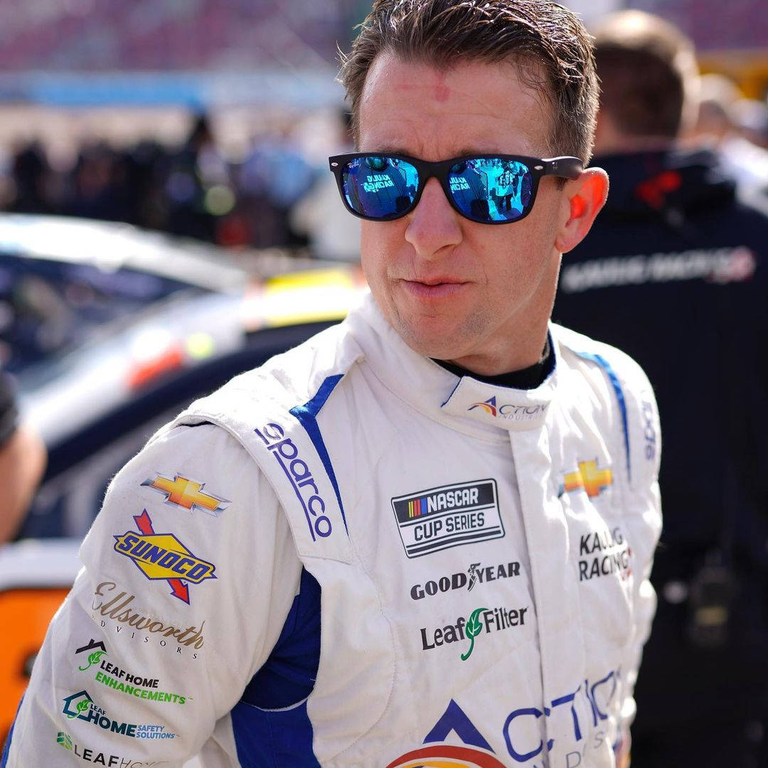 Racing 桌布 壁紙 — 一名NASCAR車手身穿白色賽車服和藍色太陽眼鏡，自信地站在賽車場上，背景是模糊的汽車和人群。