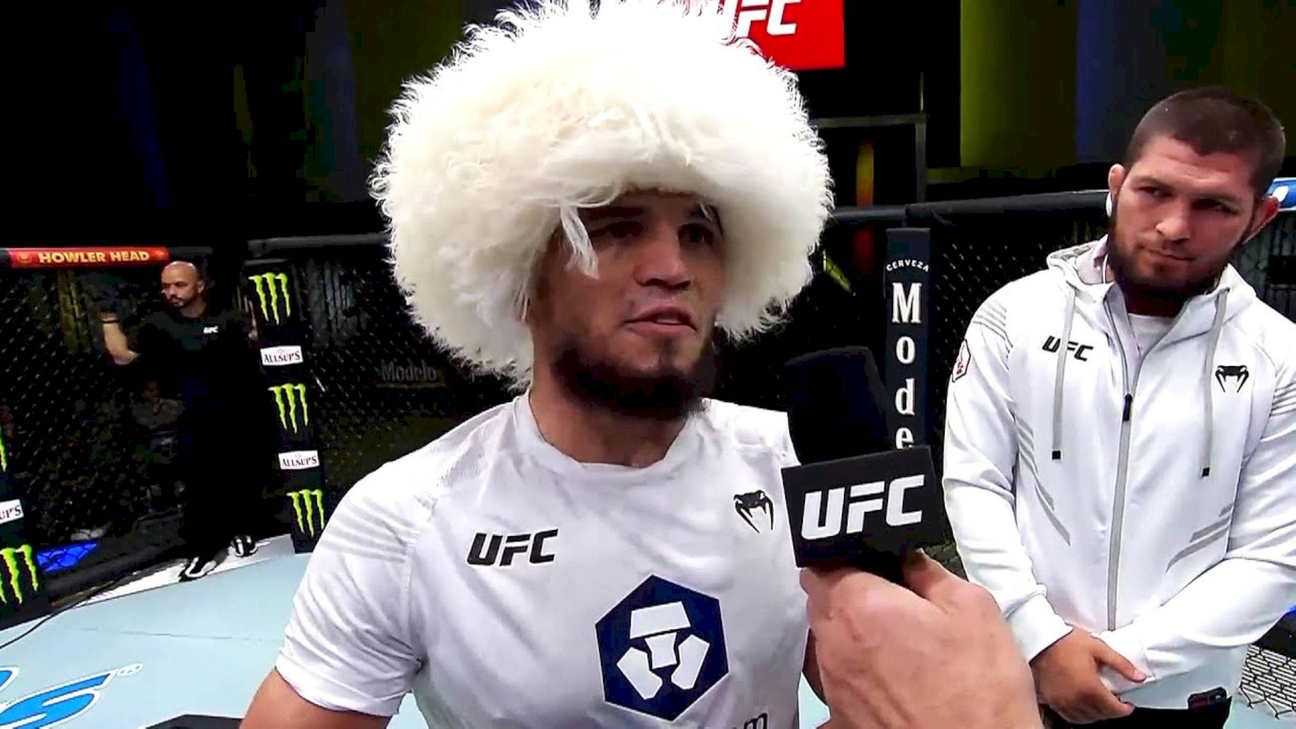 UFC 桌布 壁紙 — 一名UFC拳手戴著毛茸茸的白色帽子，正在八角籠內接受訪問，另一名拳手站在附近。