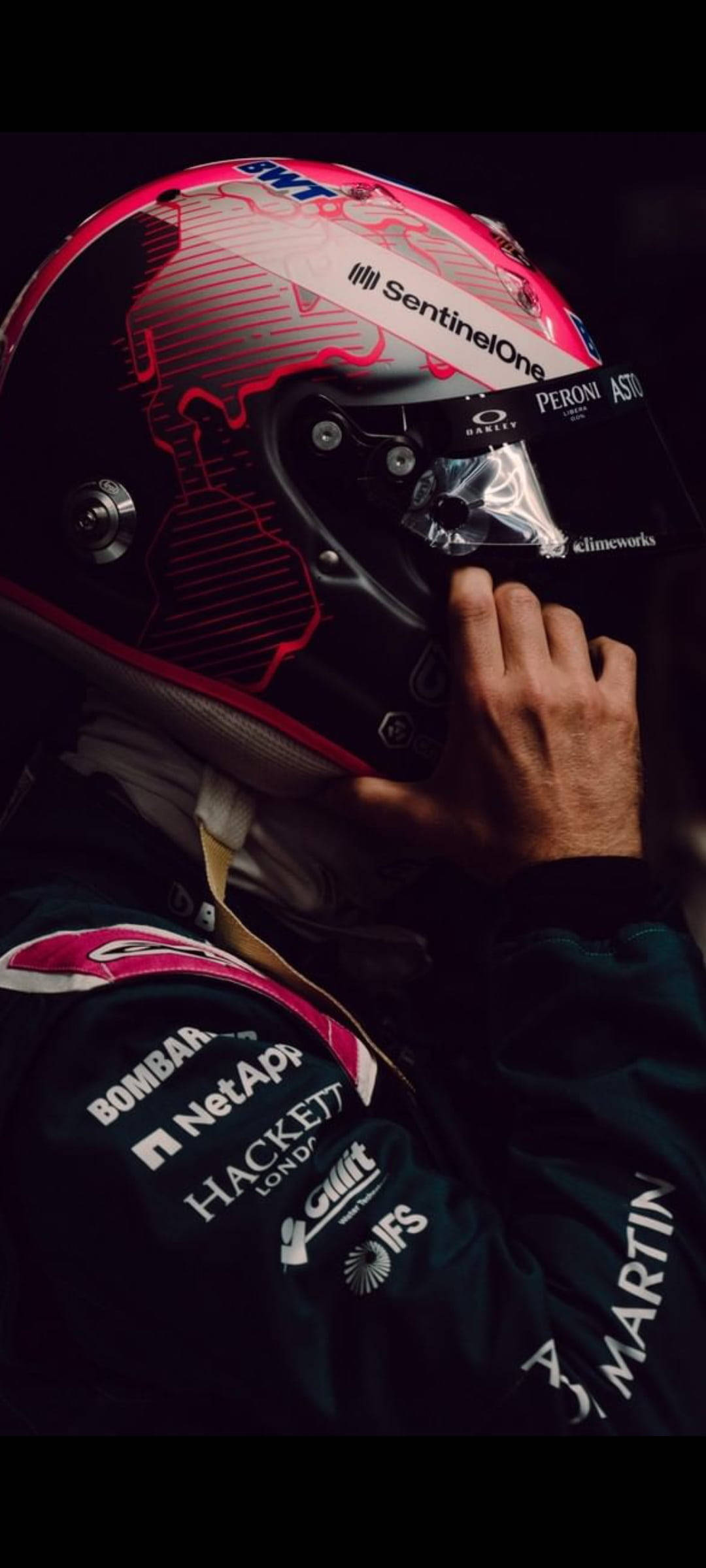 sebastian vettel 桌布 壁紙 — 一名一級方程式賽車手在戲劇性燈光下調整他帶有贊助商標誌的粉紅色和黑色頭盔。