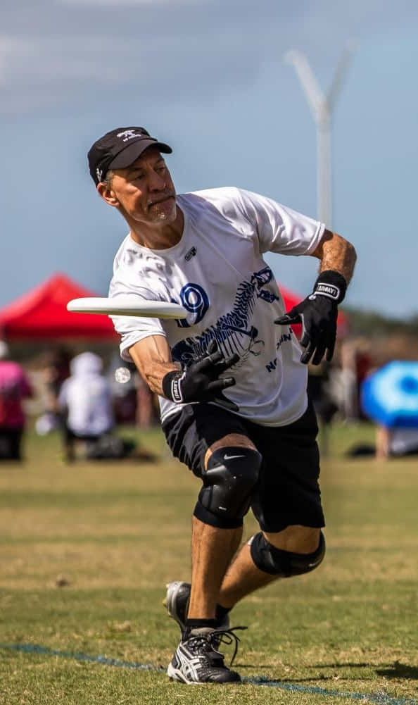 Frisbee 桌布 壁紙 — 一名中年男子穿著運動裝備，在陽光明媚的草地上向前撲去接住一個白色飛盤，背景有風力發電機。