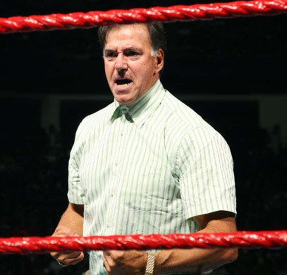 WWE 桌布 壁紙 — 一名中年男子身穿條紋襯衫，在明亮的燈光下，站在有紅色繩索的摔角擂台上，神情專注。