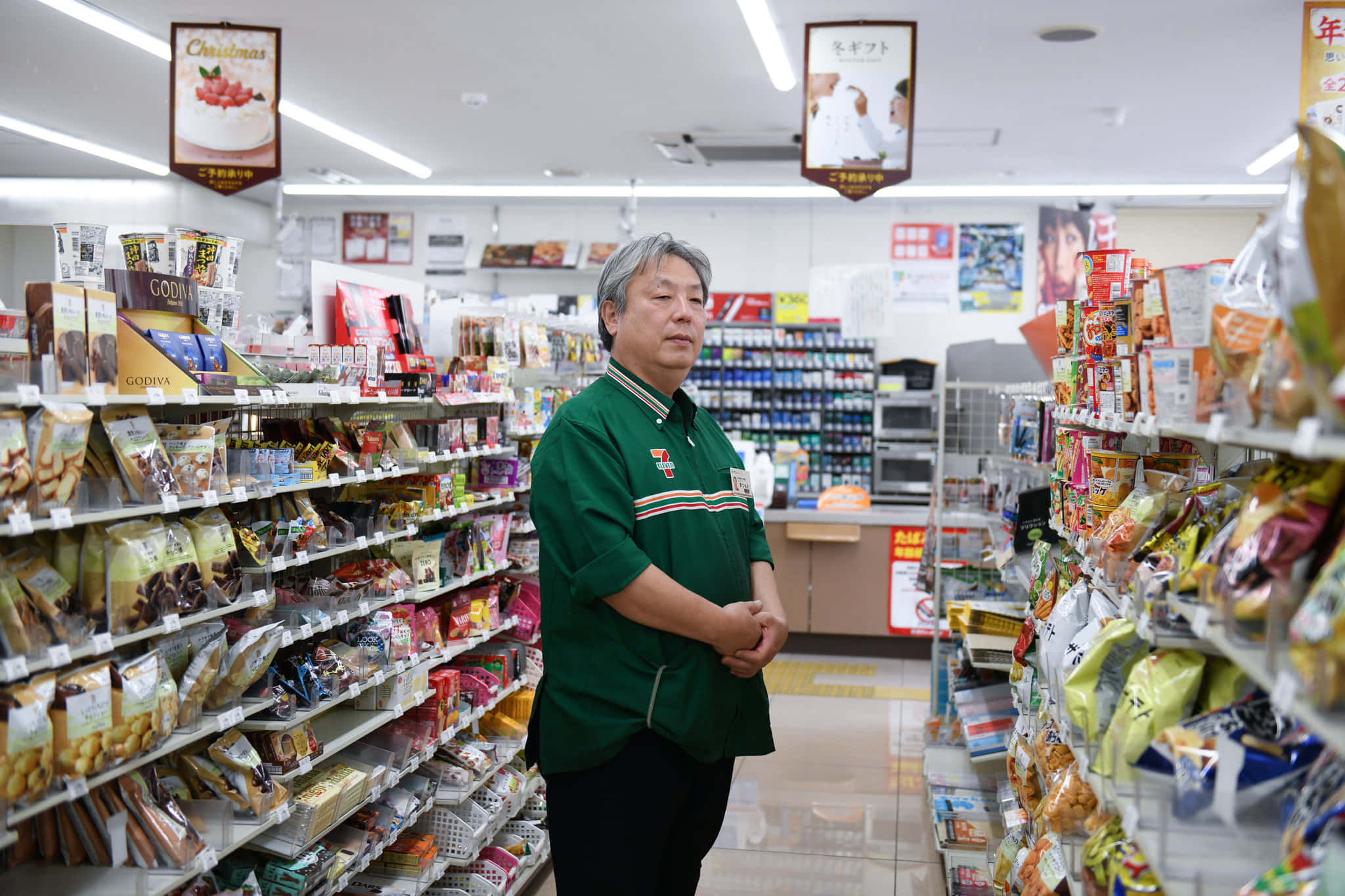 7-ELEVEN 桌布 壁紙 — 一名中年男子身穿綠色制服，平靜地站在日本便利商店明亮燈光的貨架之間，店內充滿零食和商品。