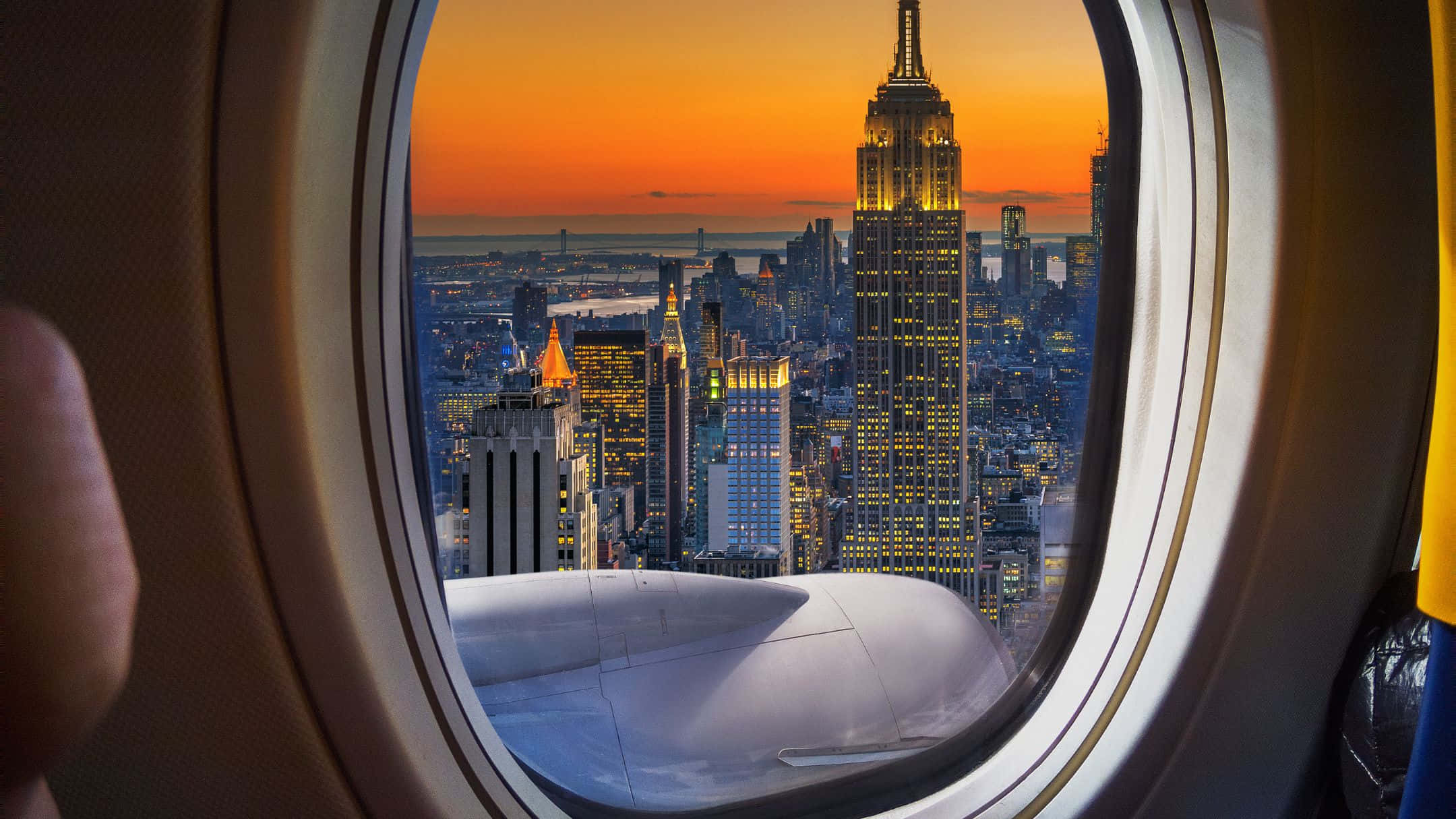 Traveling 桌布 壁紙 — 一名乘客從飛機窗戶凝視紐約市上空 vibrant 的日落，帝國大廈在金色光芒中閃耀。