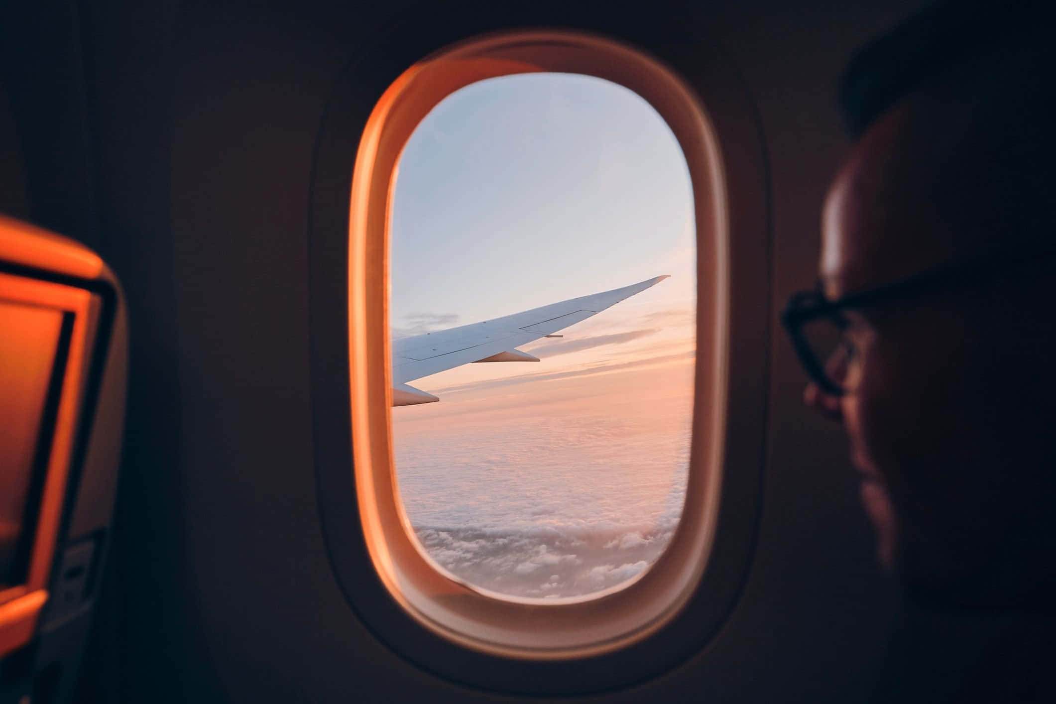 Airplane Window 桌布 壁紙 — 一名乘客從飛機窗戶向外凝視，看著機翼劃過沐浴在溫暖日落色調中的雲層。