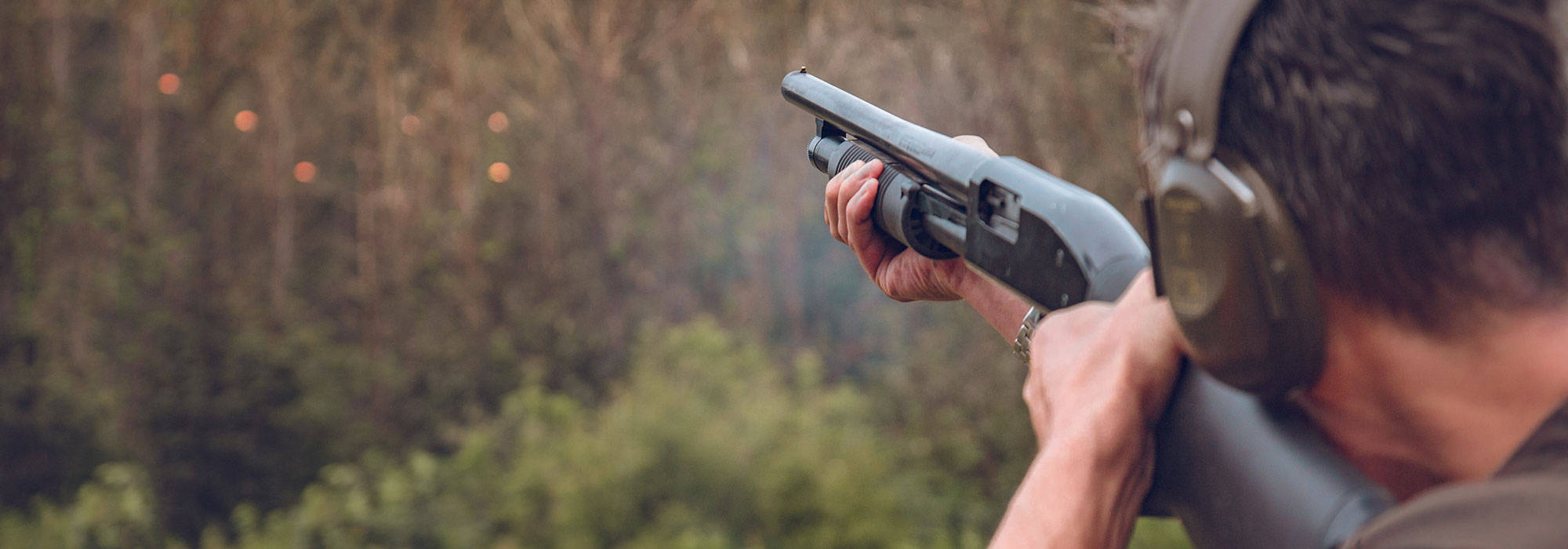 Shooting 桌布 壁紙 — 一名佩戴聽力保護裝置的人士在樹林環繞的戶外射擊場用霰彈槍瞄準目標。