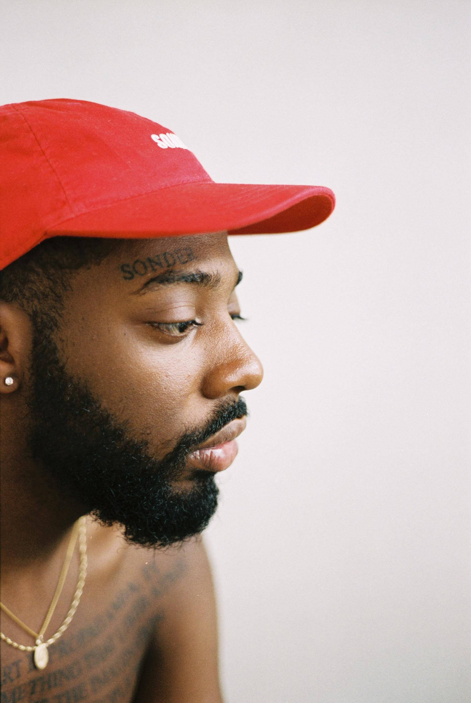 Brent Faiyaz 桌布 壁紙 — 一名側面男子戴著紅色帽子和金鏈，背景為純白色，臉部和胸部有紋身可見。