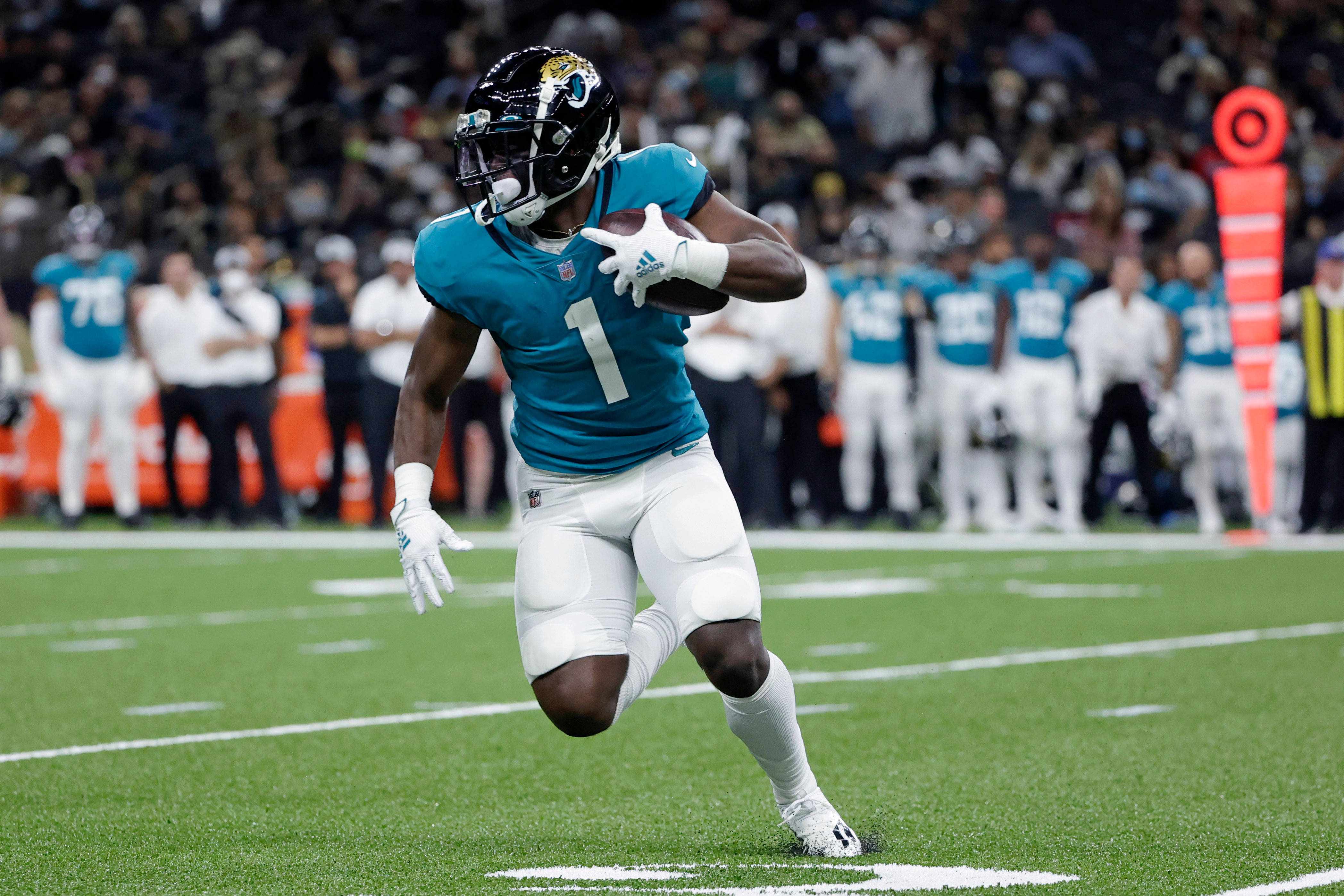 Travis Etienne 桌布 壁紙 — 一名傑克遜維爾美洲虎隊的美式足球員，身穿青色球衣一號，在比賽中帶球衝過綠色球場。