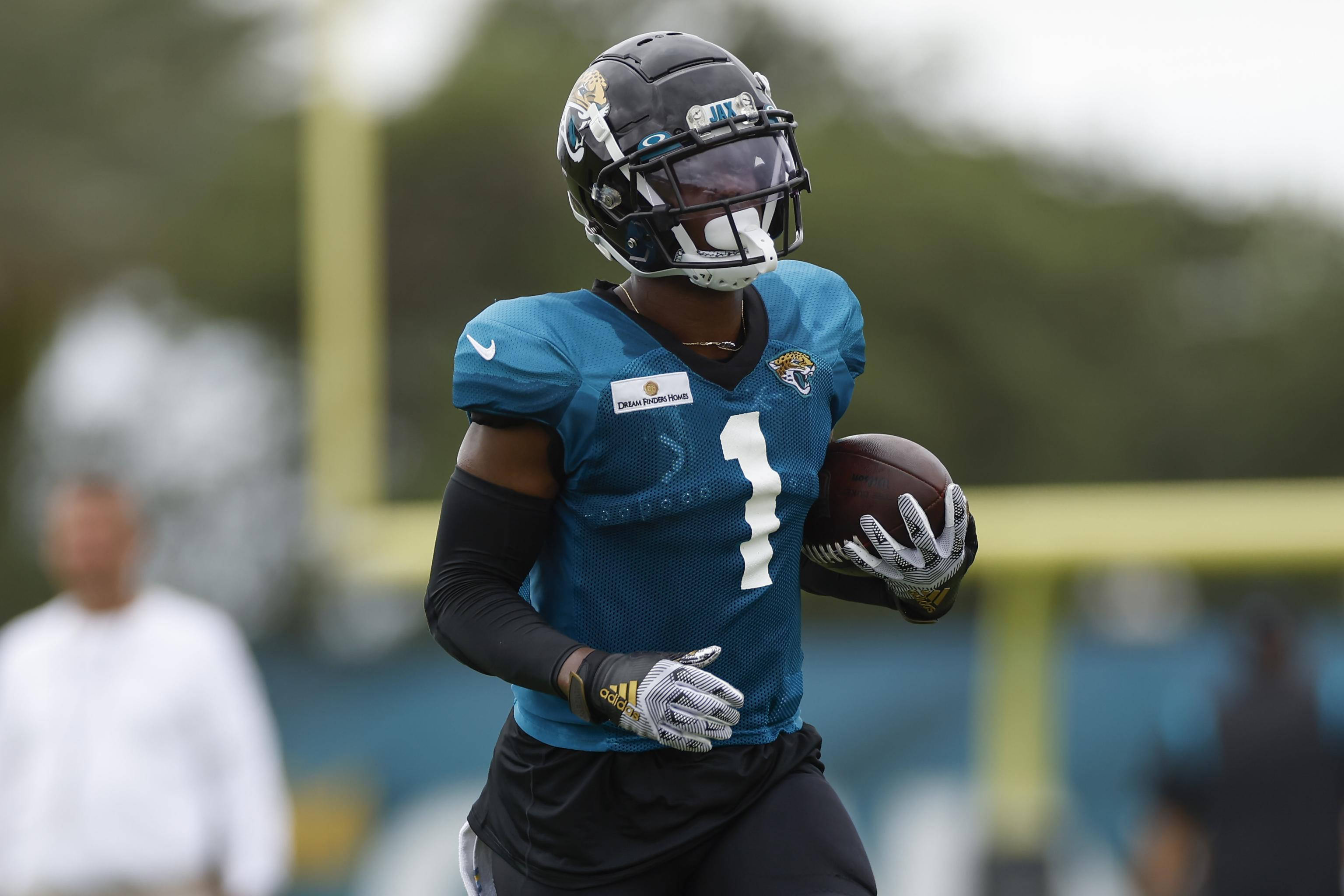 Travis Etienne 桌布 壁紙 — 一名傑克遜維爾美洲虎隊的足球員，身穿藍色一號球衣，在陰天練習時帶著足球奔跑。
