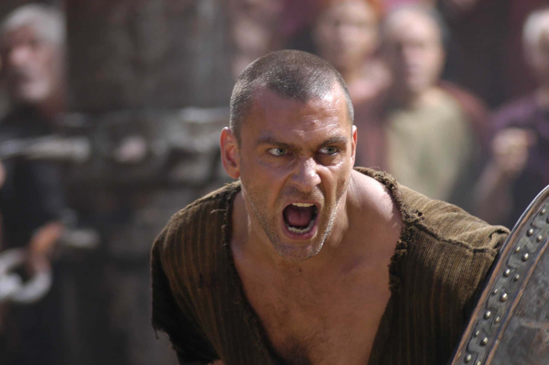 Ray Stevenson 桌布 壁紙 — 一名兇猛的角鬥士在擁擠的競技場中緊握盾牌，發出強烈的怒吼。