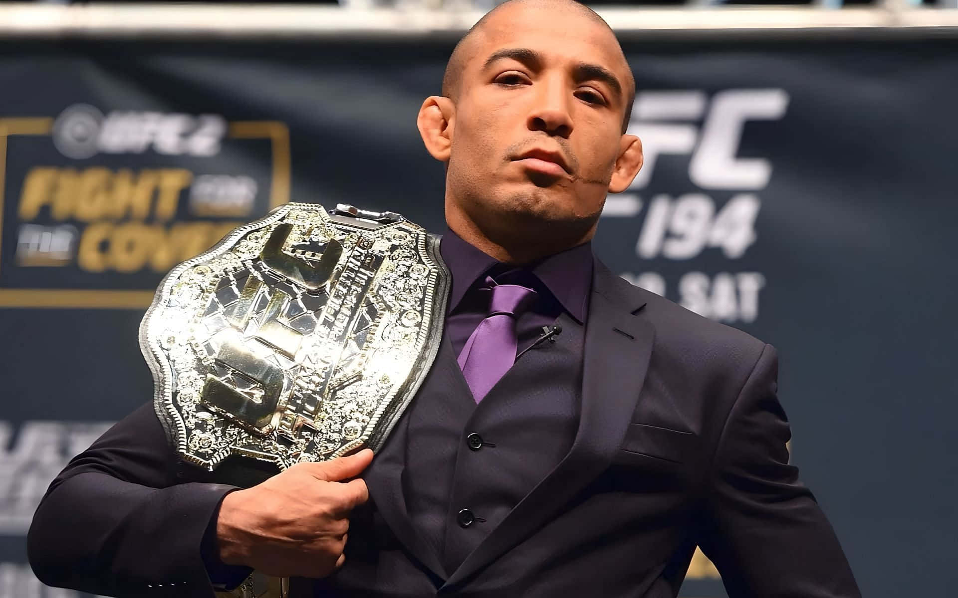 UFC 桌布 壁紙 — 一名光頭UFC拳手身穿深色西裝，在品牌活動背景前手持閃閃發光的冠軍腰帶。