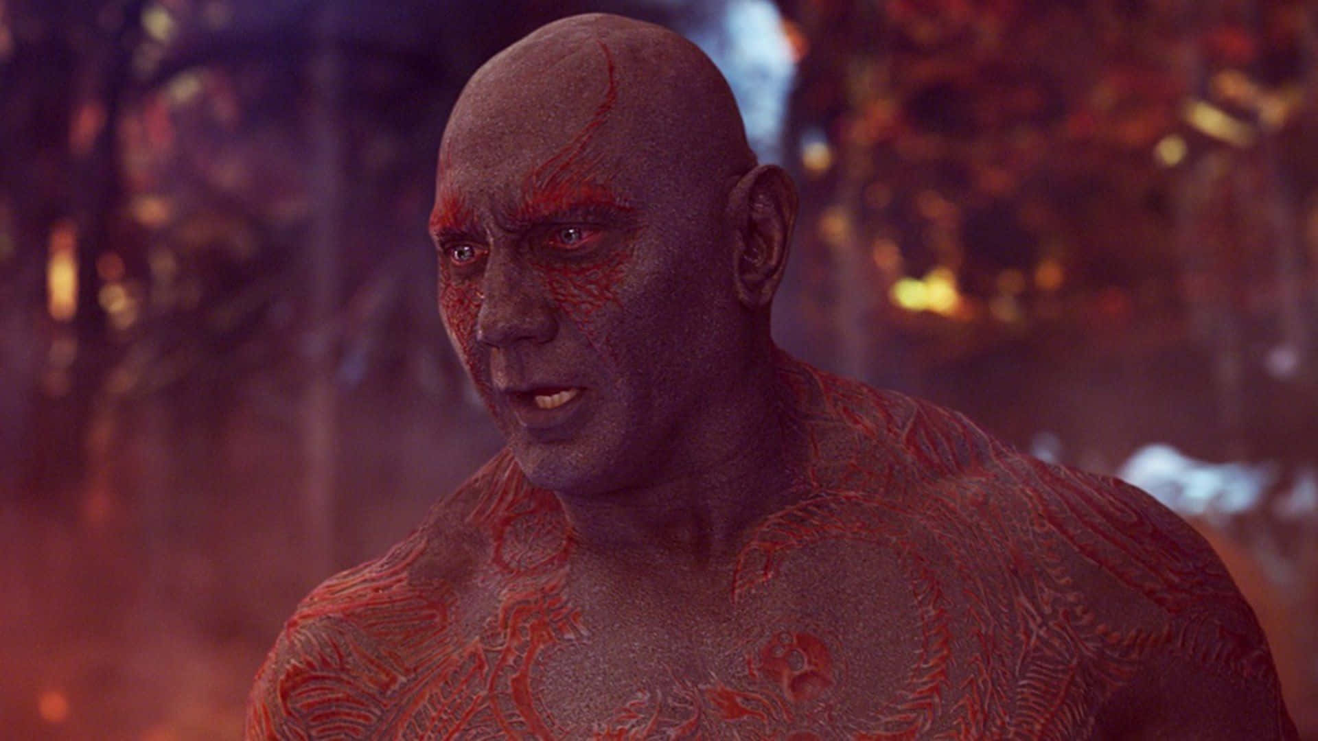 Drax 桌布 壁紙 — 一名光頭男子，臉上和軀幹上有紅色部落圖案，站在充滿火焰和煙霧的環境中，背景有模糊的樹木。