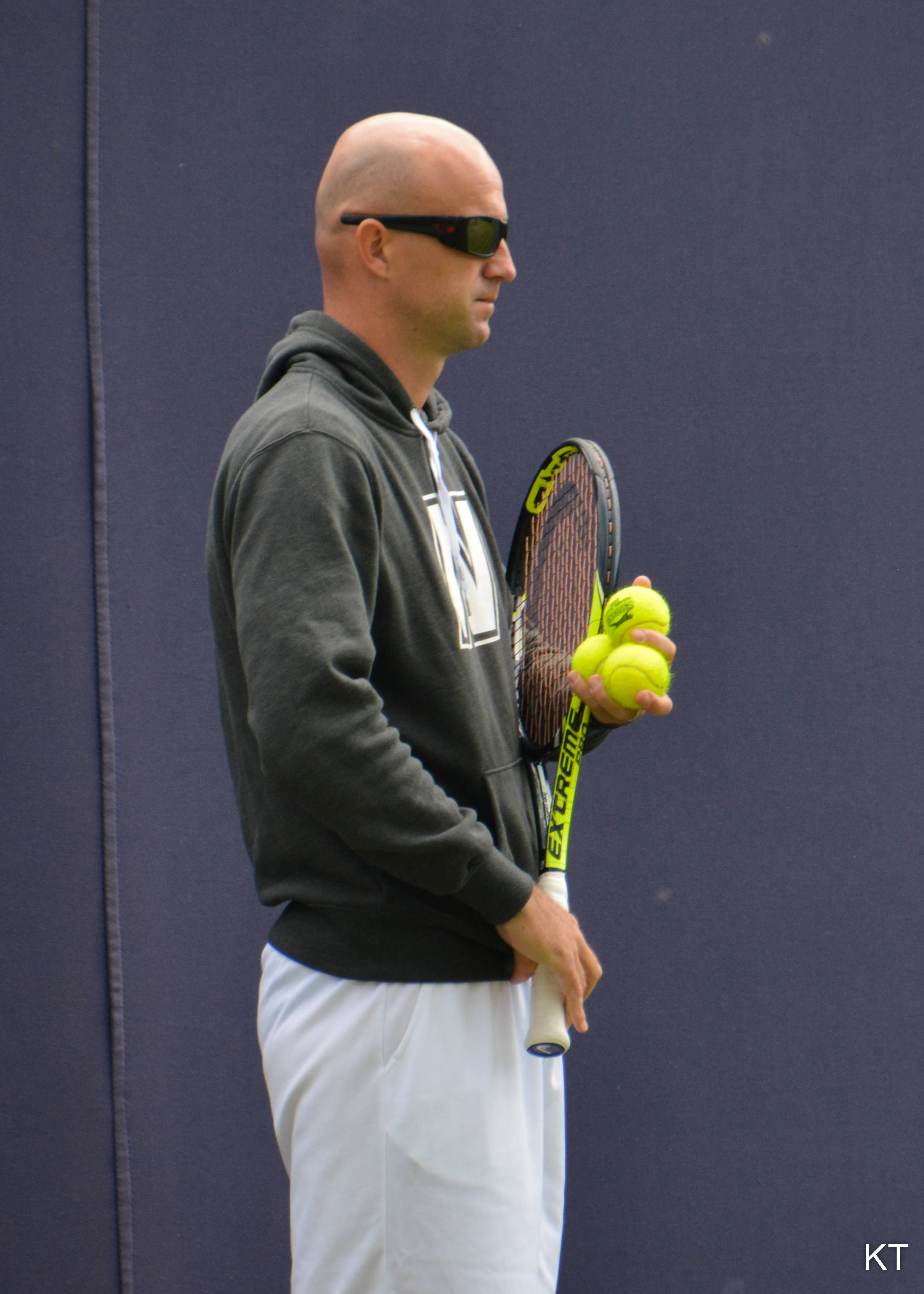 Tennis 桌布 壁紙 — 一名光頭男子戴著太陽眼鏡，手持網球拍和亮黃色網球，背景是深藍色牆壁。