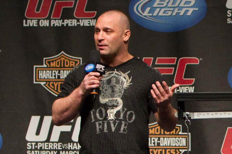 MMA 桌布 壁紙 — 一名光頭男子穿著圖案T恤，在UFC記者會上對著麥克風講話，身後有贊助商標誌。