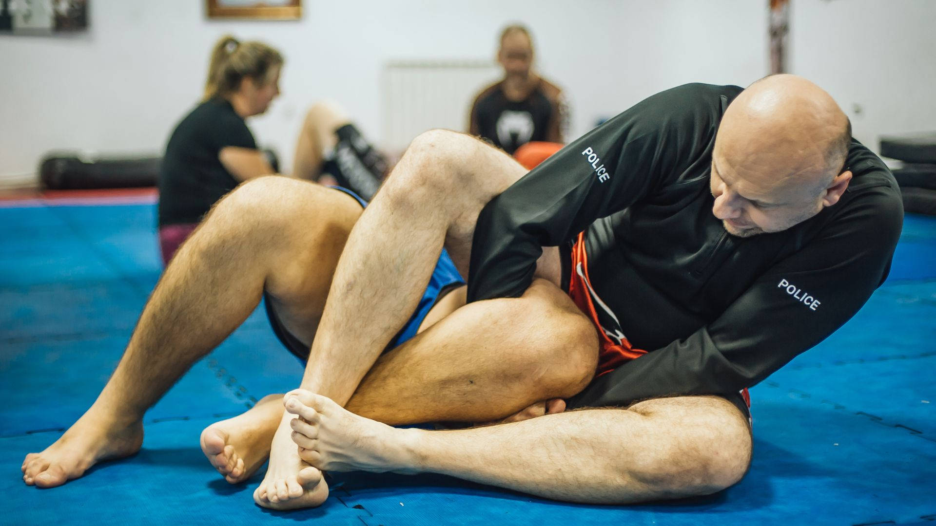 martial arts 桌布 壁紙 — 一名光頭男子穿著黑色警察柔道服，在藍色墊子上執行纏鬥技術，背景中其他人正在訓練。