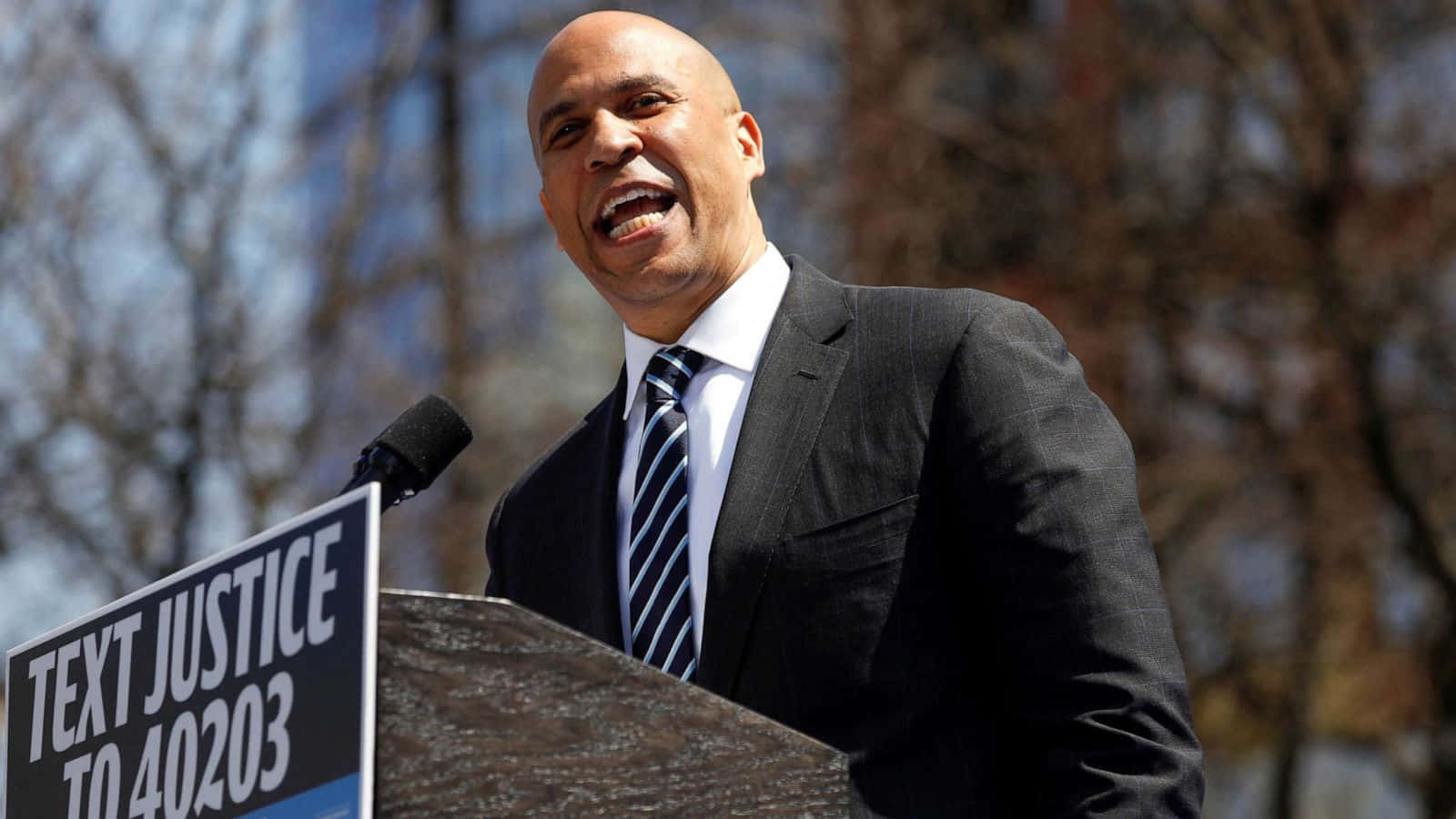 MacBook 動態圖片 桌布 壁紙 — 一名光頭男子身穿深色西裝和條紋領帶，在戶外講台上對著麥克風熱情演講，旁邊有『TEXT JUSTICE TO 40203』的標誌。