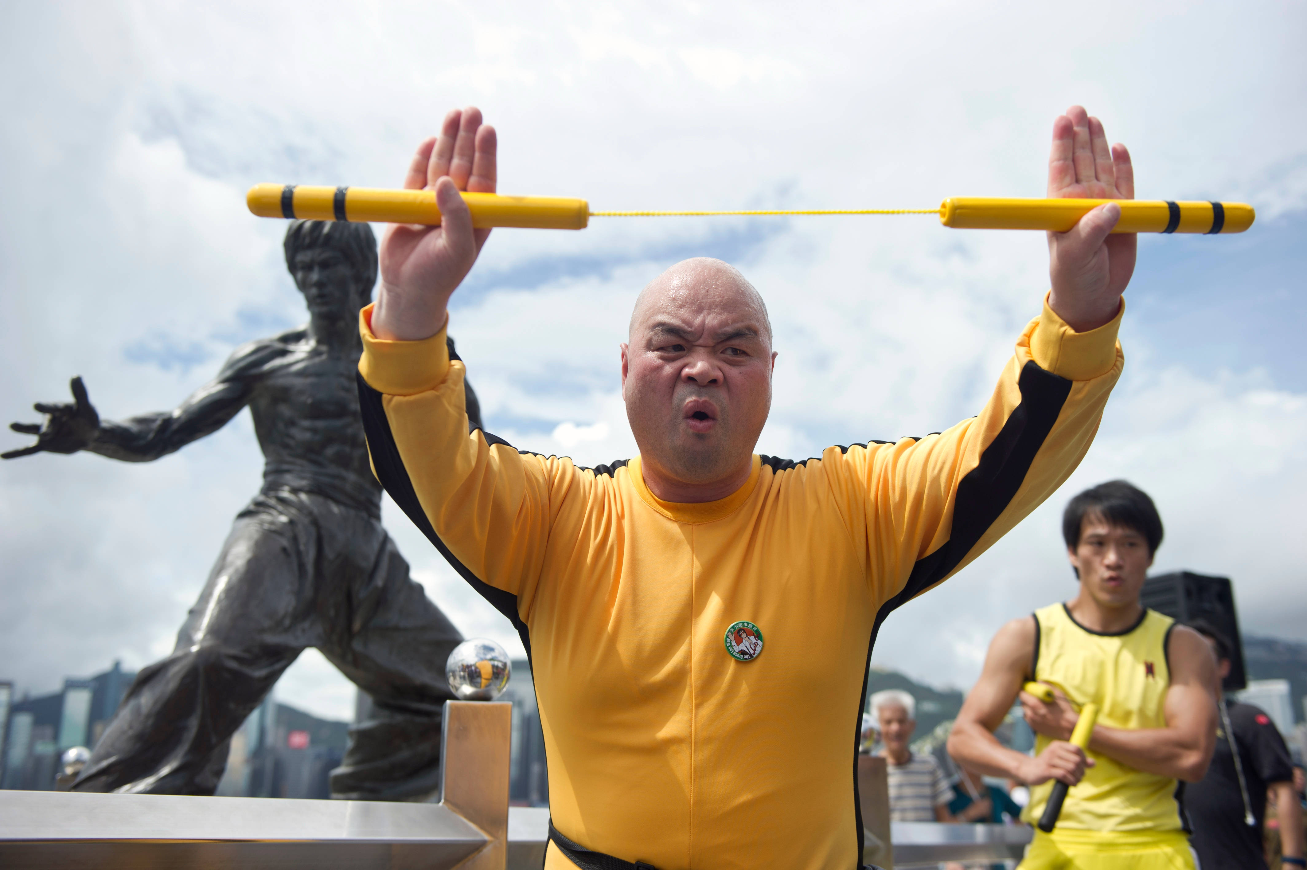 Exercise 桌布 壁紙 — 一名光頭男子身穿黃色武術制服，在李小龍雕像和城市天際線的背景下，以專注的神情擺出動態的雙節棍姿勢。