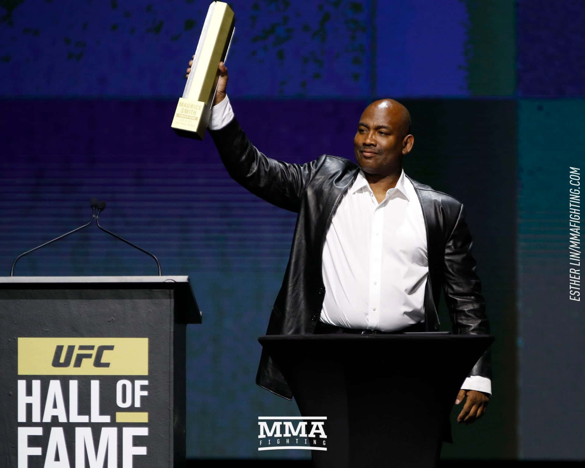 MMA 桌布 壁紙 — 一名光頭男子身穿黑色皮夾克，在明亮燈光下於舞台上舉起金色UFC名人堂獎盃。