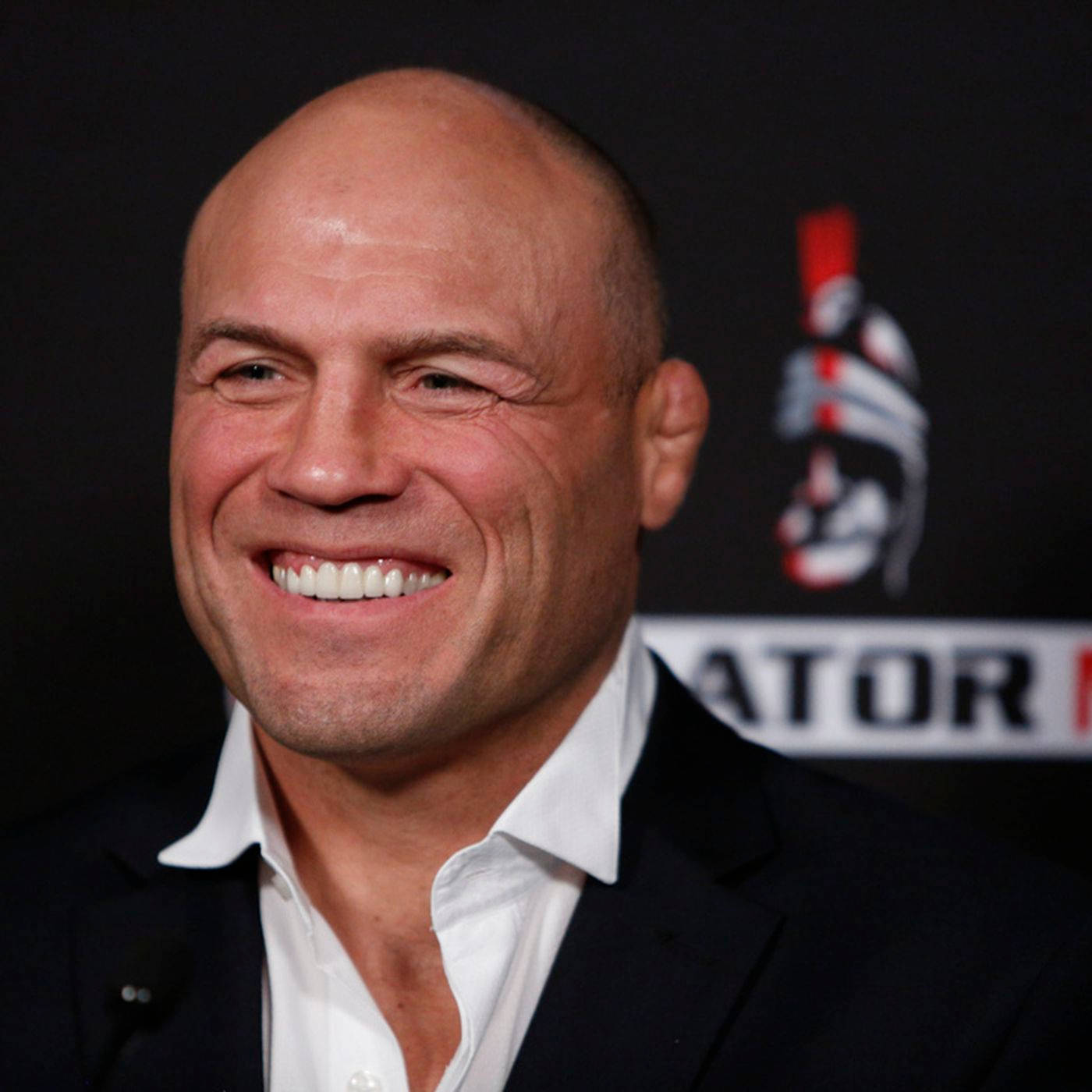MMA 桌布 壁紙 — 一名光頭男子身穿黑色西裝和白色襯衫，在帶有標誌的深色背景的記者會上燦爛地微笑。