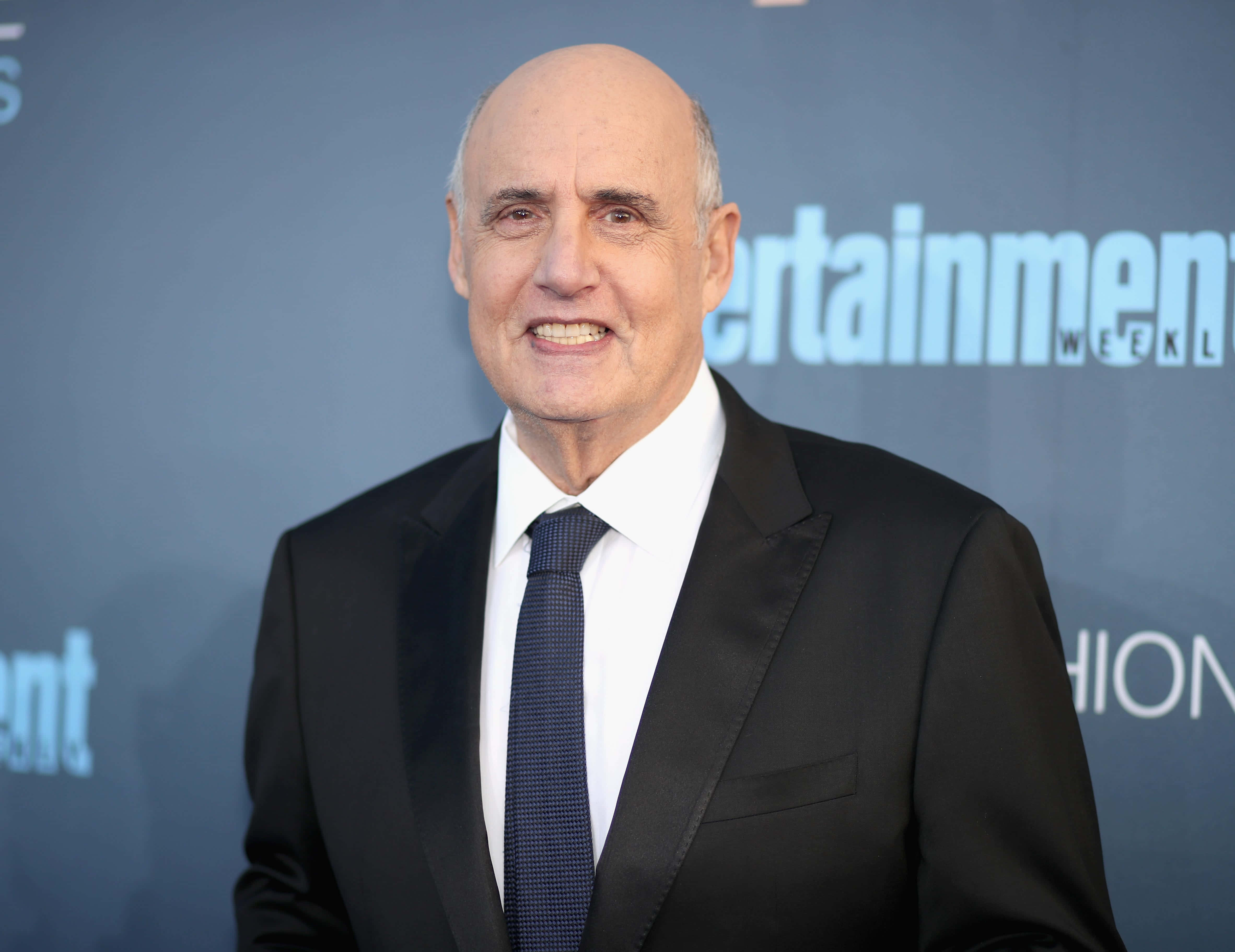 jeffrey tambor 桌布 壁紙 — 一名光頭男子身穿黑色西裝和藍色領帶，在藍色Entertainment Weekly背景前對著鏡頭微笑。