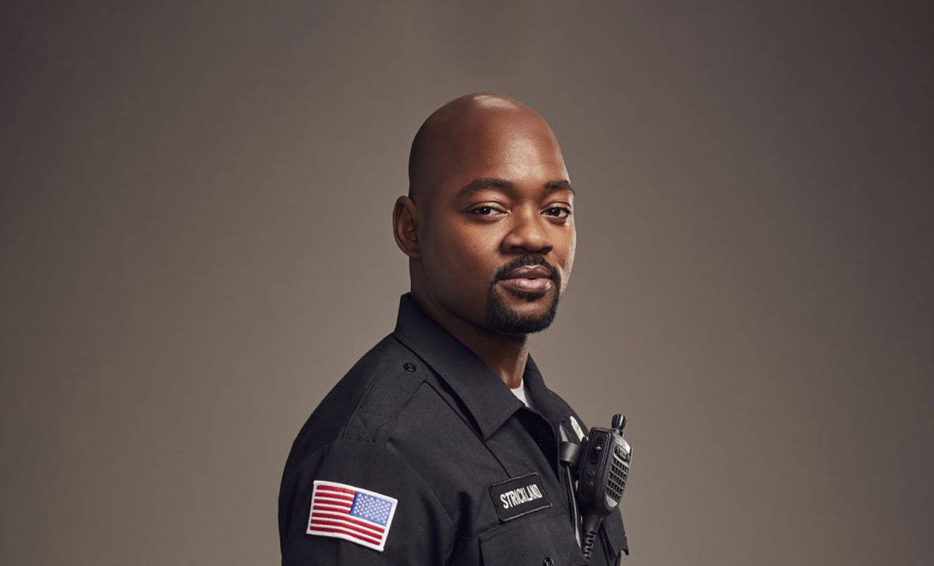Black Man 桌布 壁紙 — 一名光頭黑人男子身穿深色警察制服，帶有美國國旗臂章和對講機，在柔和的灰色背景下平靜地凝視。