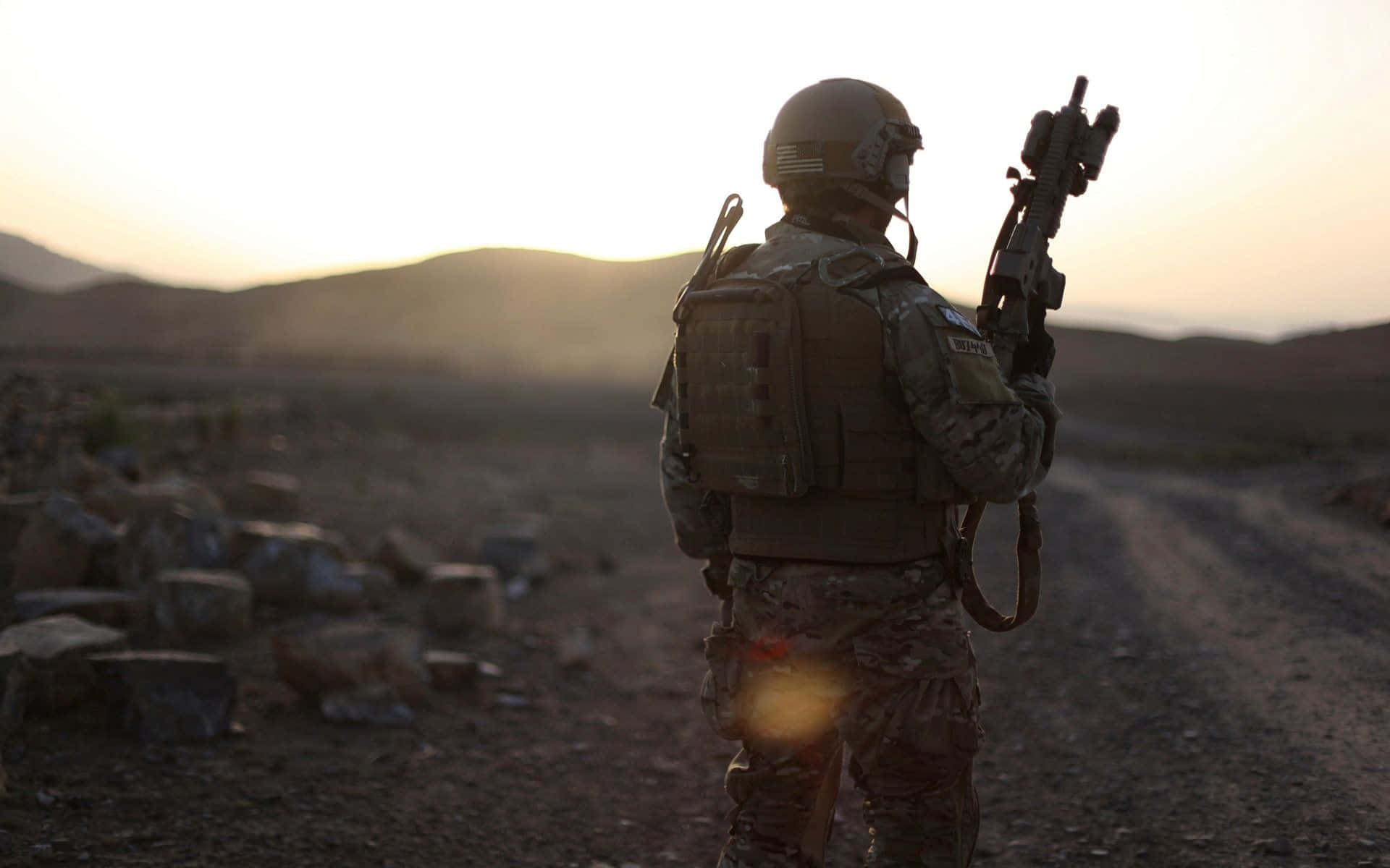  壁紙 — 一名全副武裝的士兵在日落時獨自站在塵土飛揚的道路上，剪影映襯著起伏的山丘和散落的岩石。