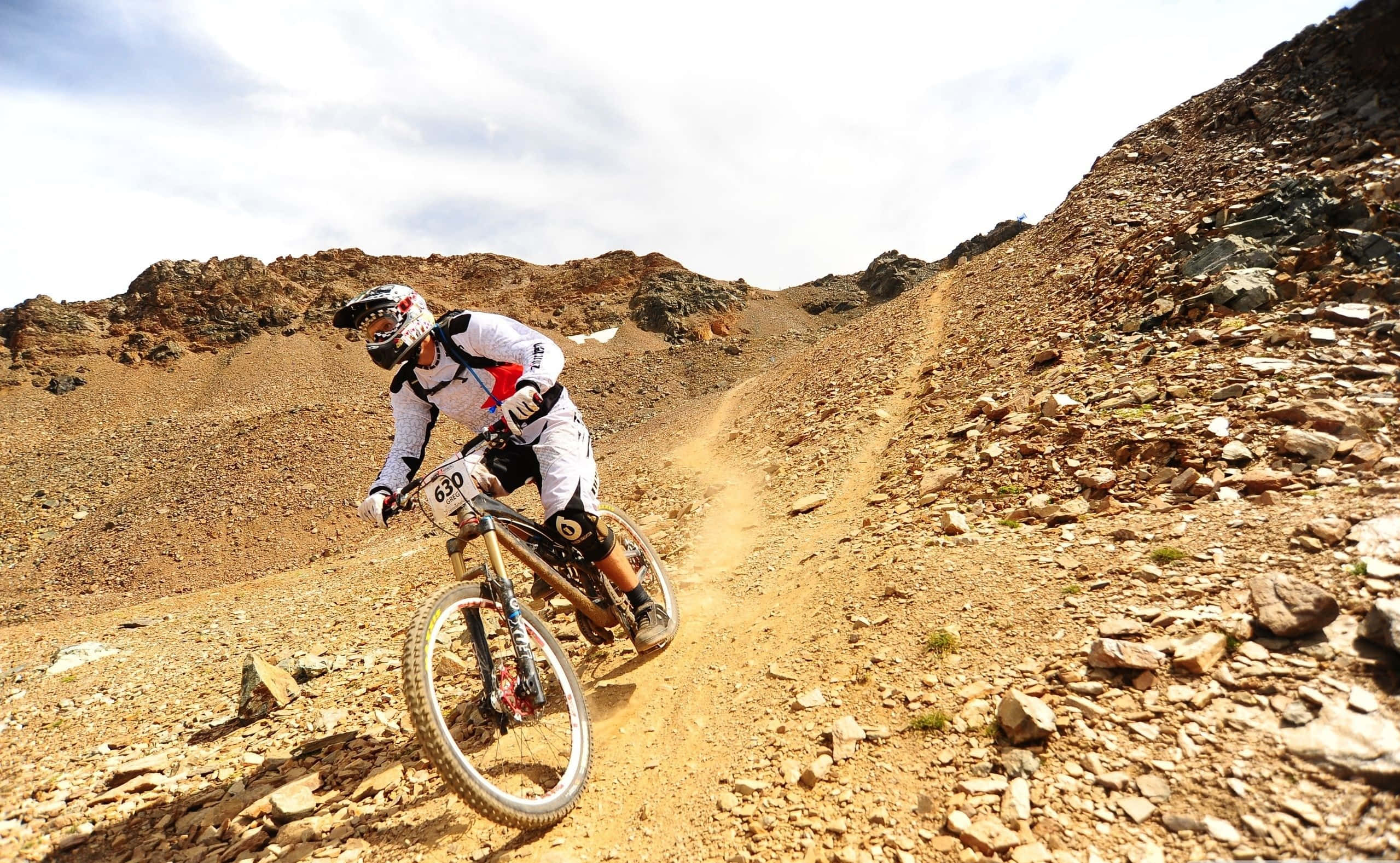 Mtb 桌布 壁紙 — 一名全副武裝的山地車手在晴朗的天空下，沿著一條崎嶇多塵的小徑，穿越岩石乾燥的地形飛馳而下。