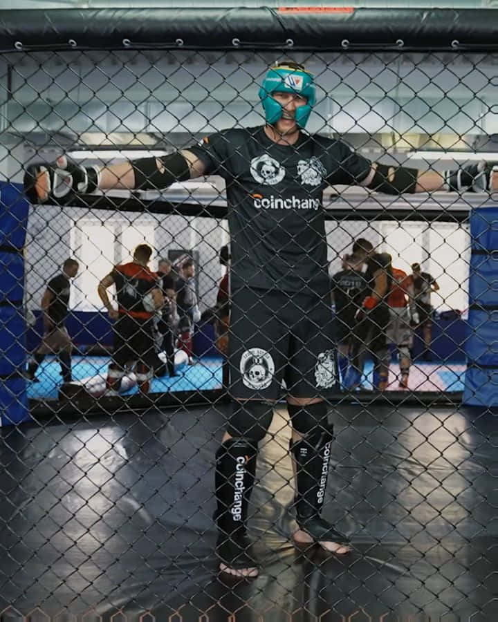 UFC 桌布 壁紙 — 一名全副武裝的戰士自信地站在MMA鐵籠內，背景有其他人在訓練。