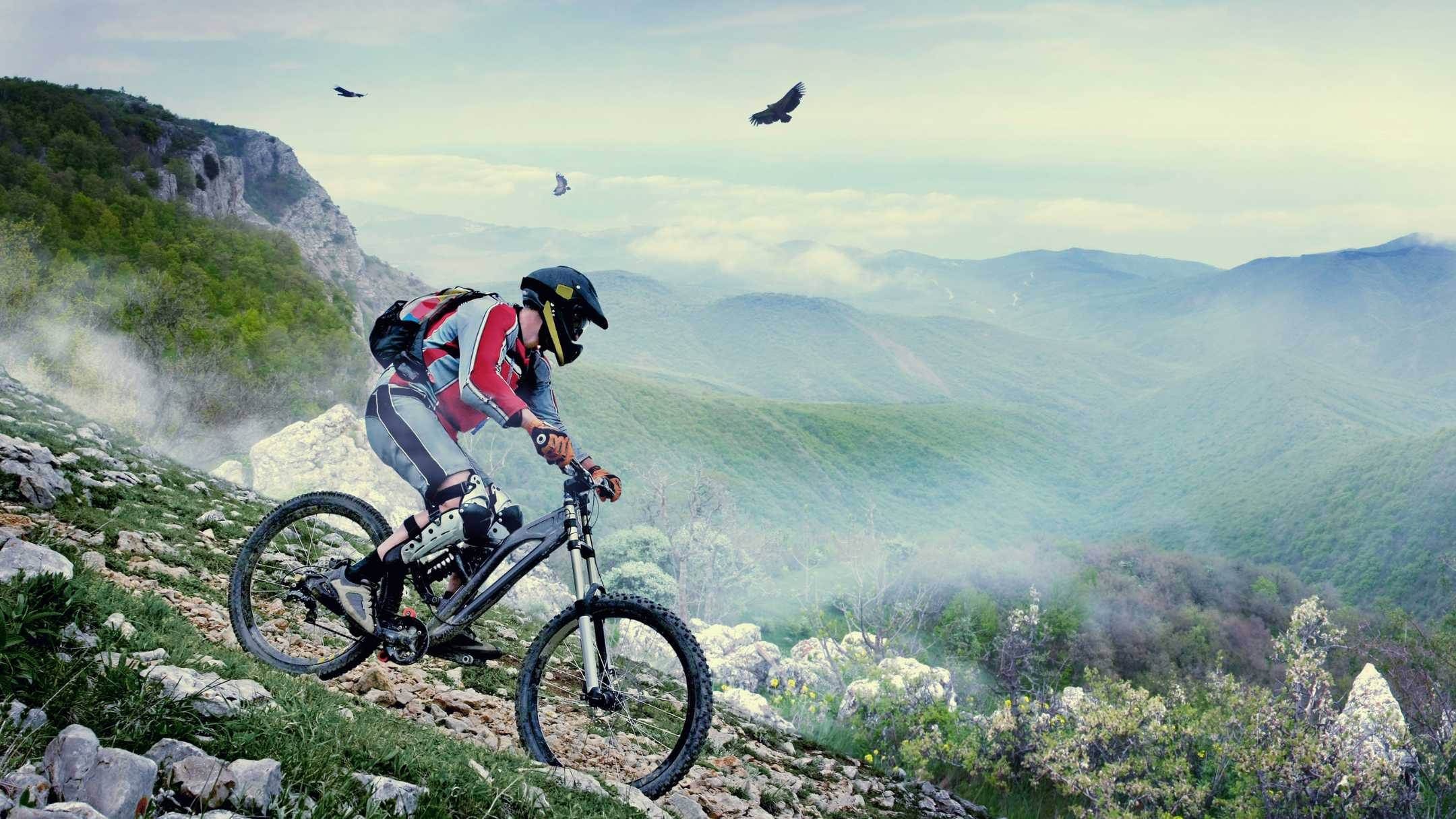 Mountain Bike 桌布 壁紙 — 一名全副裝備的山地自行車手在霧氣繚繞的山脊上沿著岩石小徑下行，頭頂有鳥兒翱翔。
