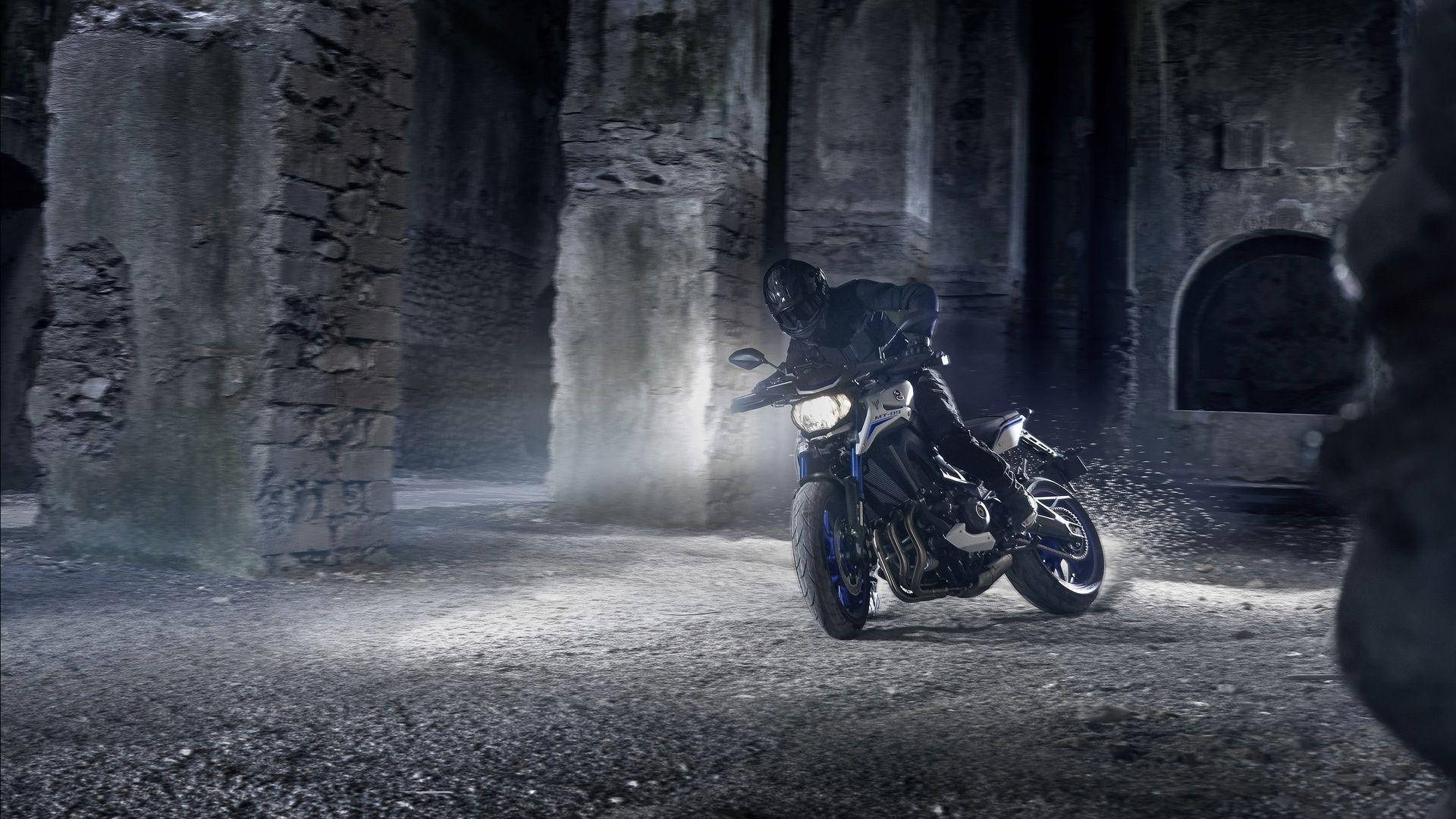 yamaha mt 15 桌布 壁紙 — 一名全副裝備的摩托車手在夜間騎著堅固的摩托車，穿過霧氣籠罩的古老石廢墟，傾身入彎。