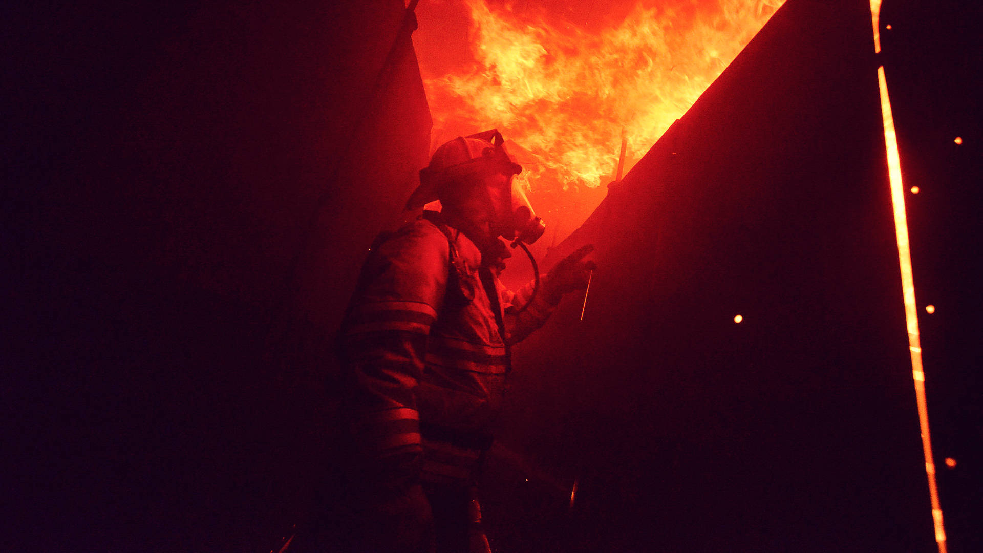 Firefighter 桌布 壁紙 — 一名全副裝備的消防員在夜間站立，身影映襯著吞噬建築物的猛烈橙色火焰。