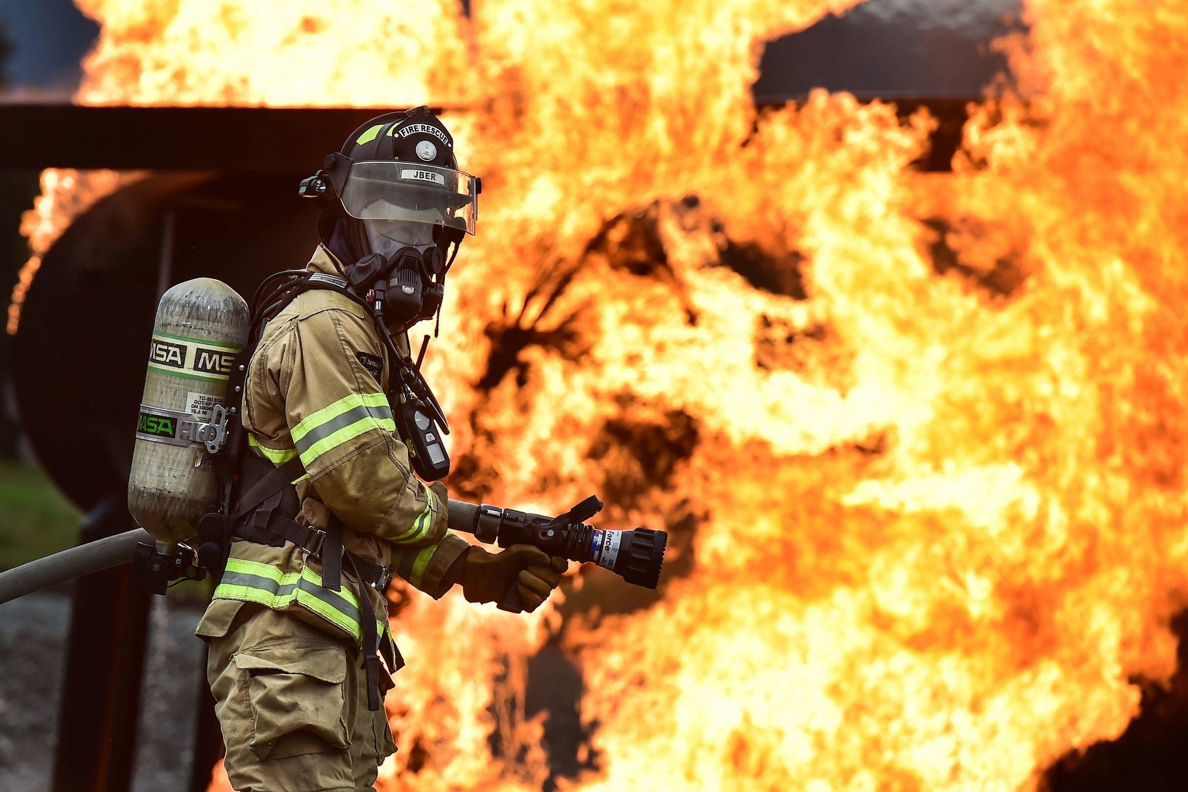 Firefighter 桌布 壁紙 — 一名全副裝備的消防員正用消防喉對抗猛烈燃燒的火焰，背景是強烈的橙色烈焰。