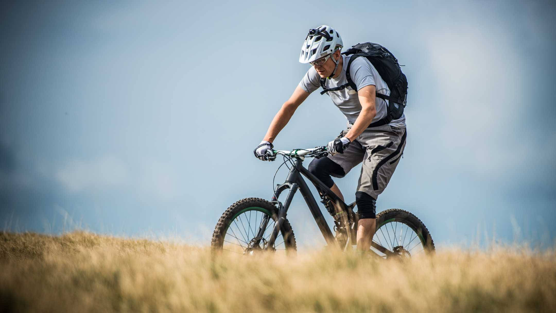 Mountain Bike 桌布 壁紙 — 一名全副裝備的自行車手在晴朗的藍天下，騎著登山自行車穿過陽光照耀的草地山坡。
