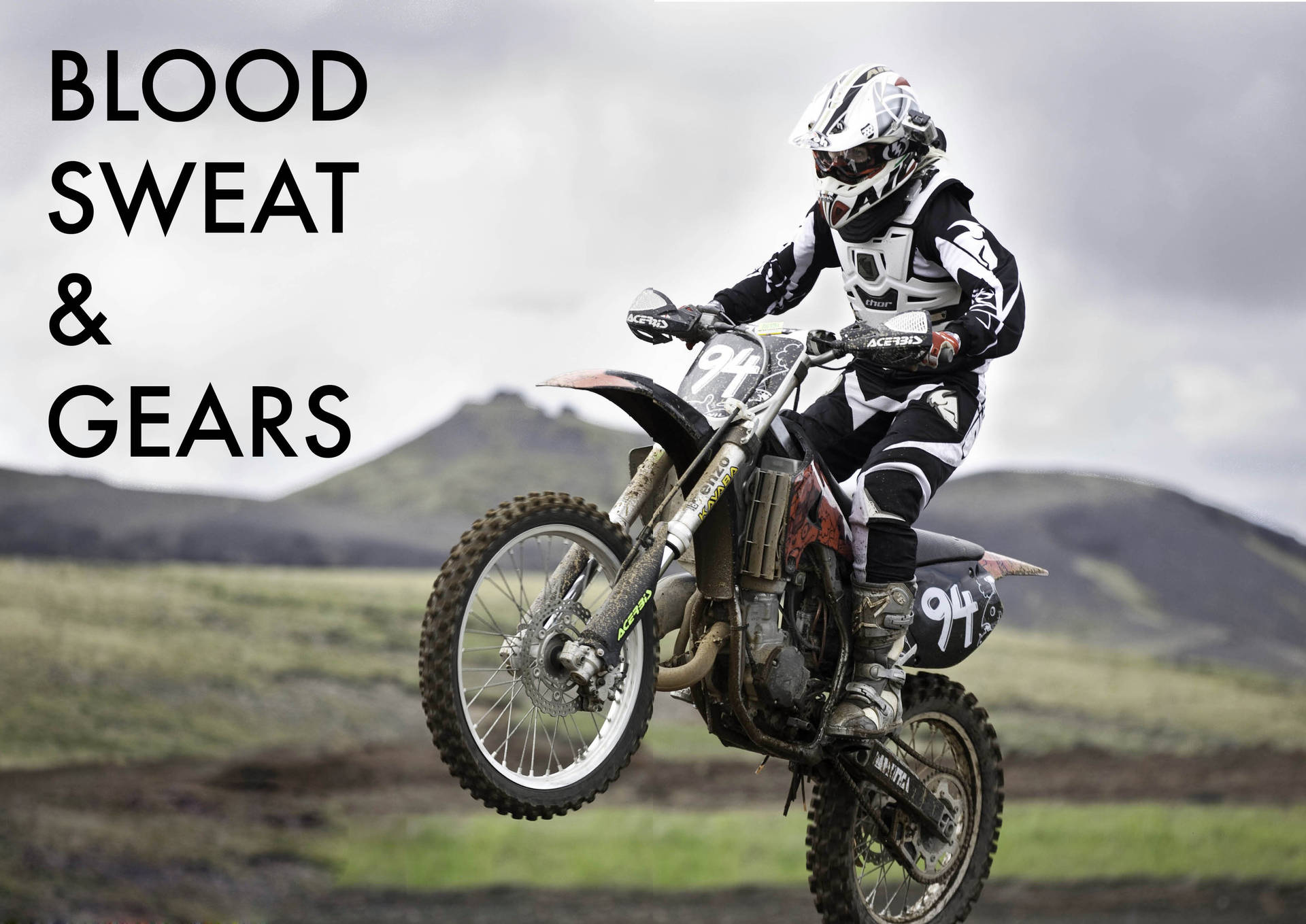 Motocross 桌布 壁紙 — 一名全副裝備的越野摩托車手在陰天之下，騎著越野摩托車飛越崎嶇的草坡，在空中翱翔。