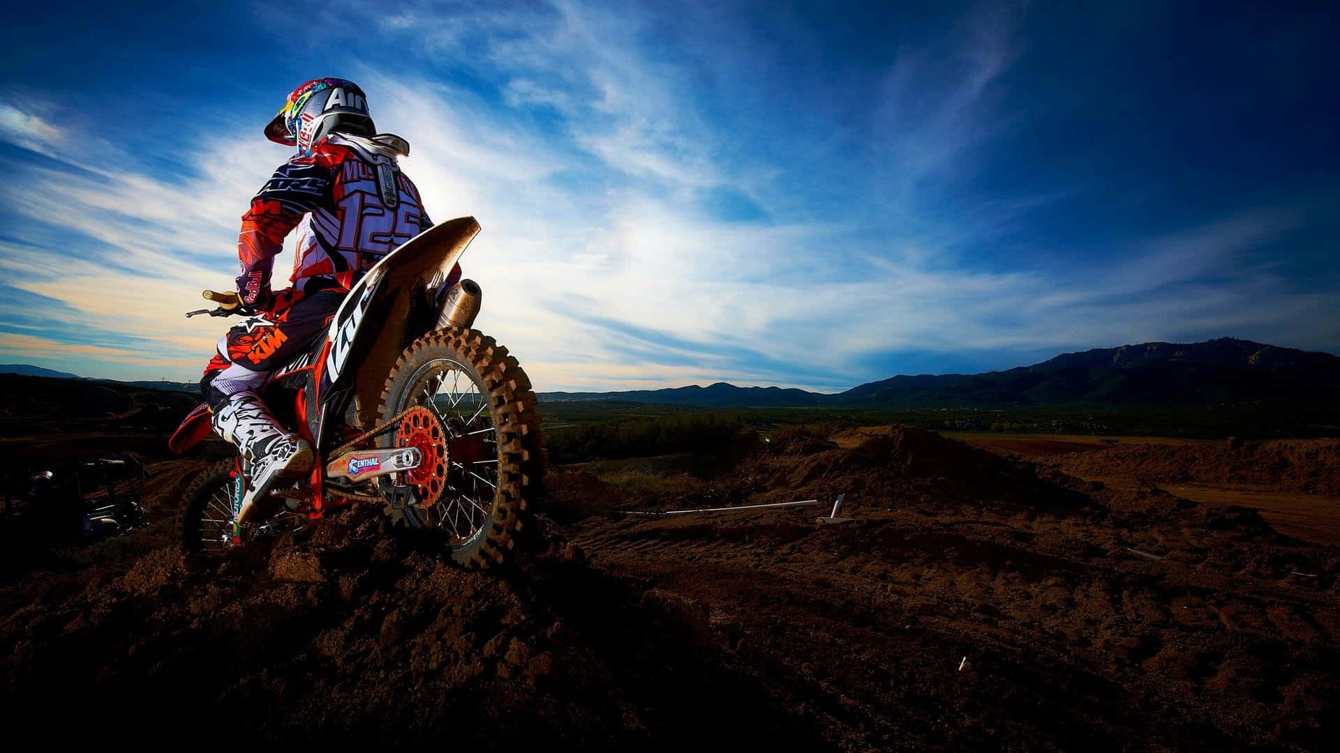 Motocross 桌布 壁紙 — 一名全副裝備的越野機車手在土丘上稍作休息，背景是戲劇性的暮色天空和遠山。